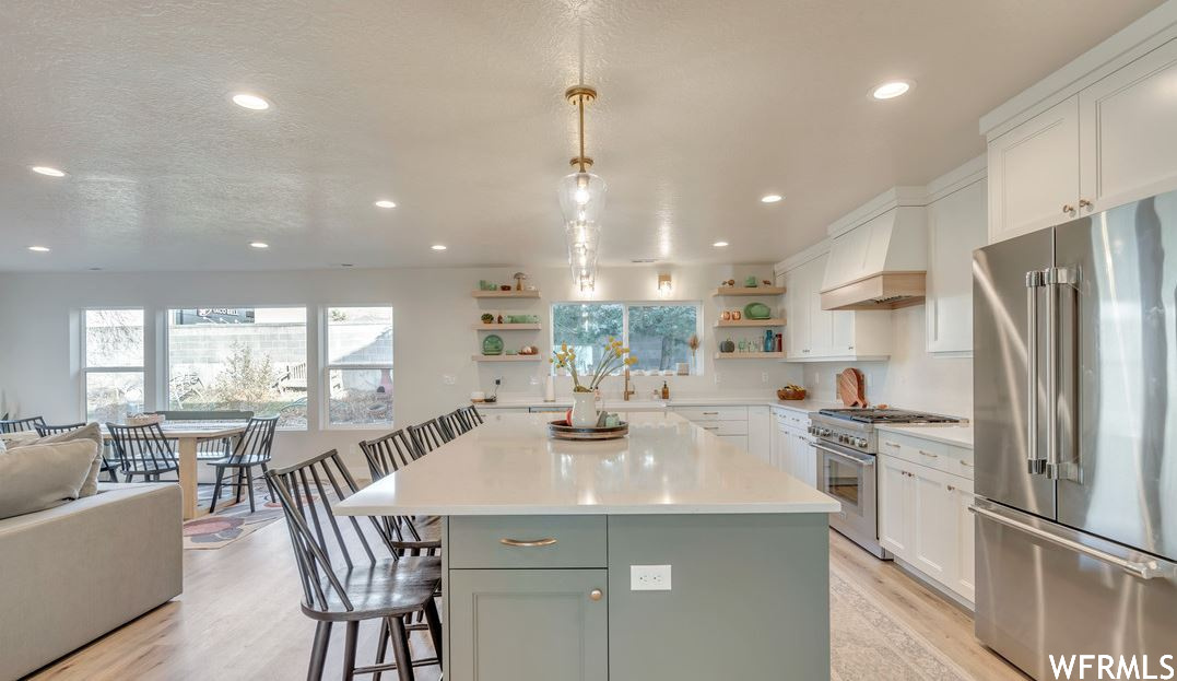 Kitchen featuring white cabinetry, a textured ceiling, a kitchen island, light countertops, light hardwood flooring, decorative light fixtures, custom range hood, a kitchen island with sink, and high end appliances