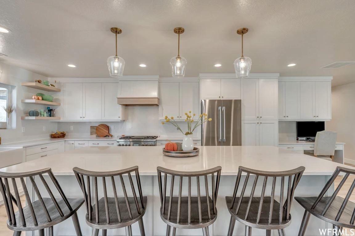 Kitchen with stainless steel appliances, a kitchen island, backsplash, light countertops, custom exhaust hood, white cabinetry, and pendant lighting