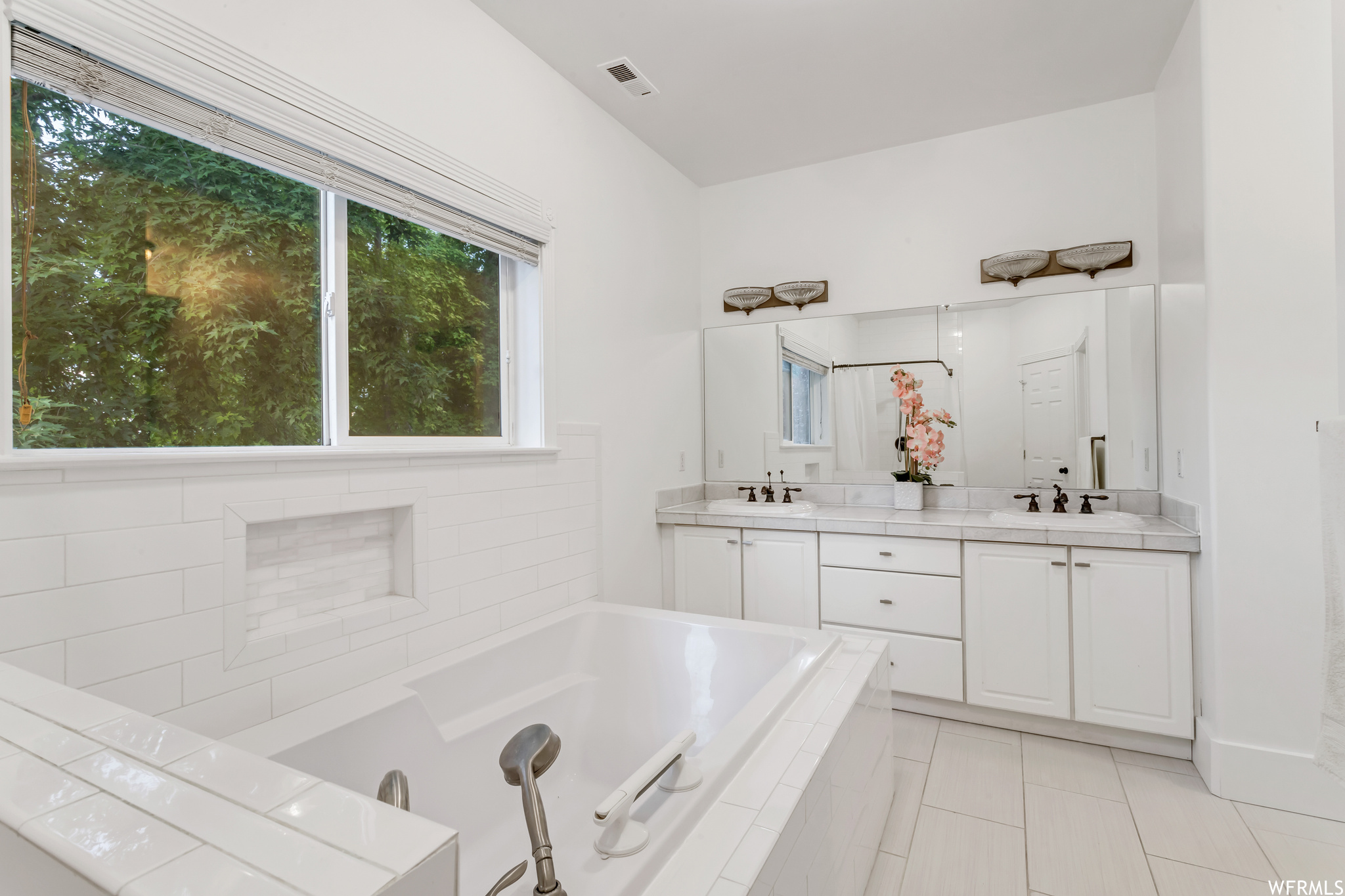Bathroom with dual bowl vanity, tiled bath, light tile floors, plenty of natural light, and mirror