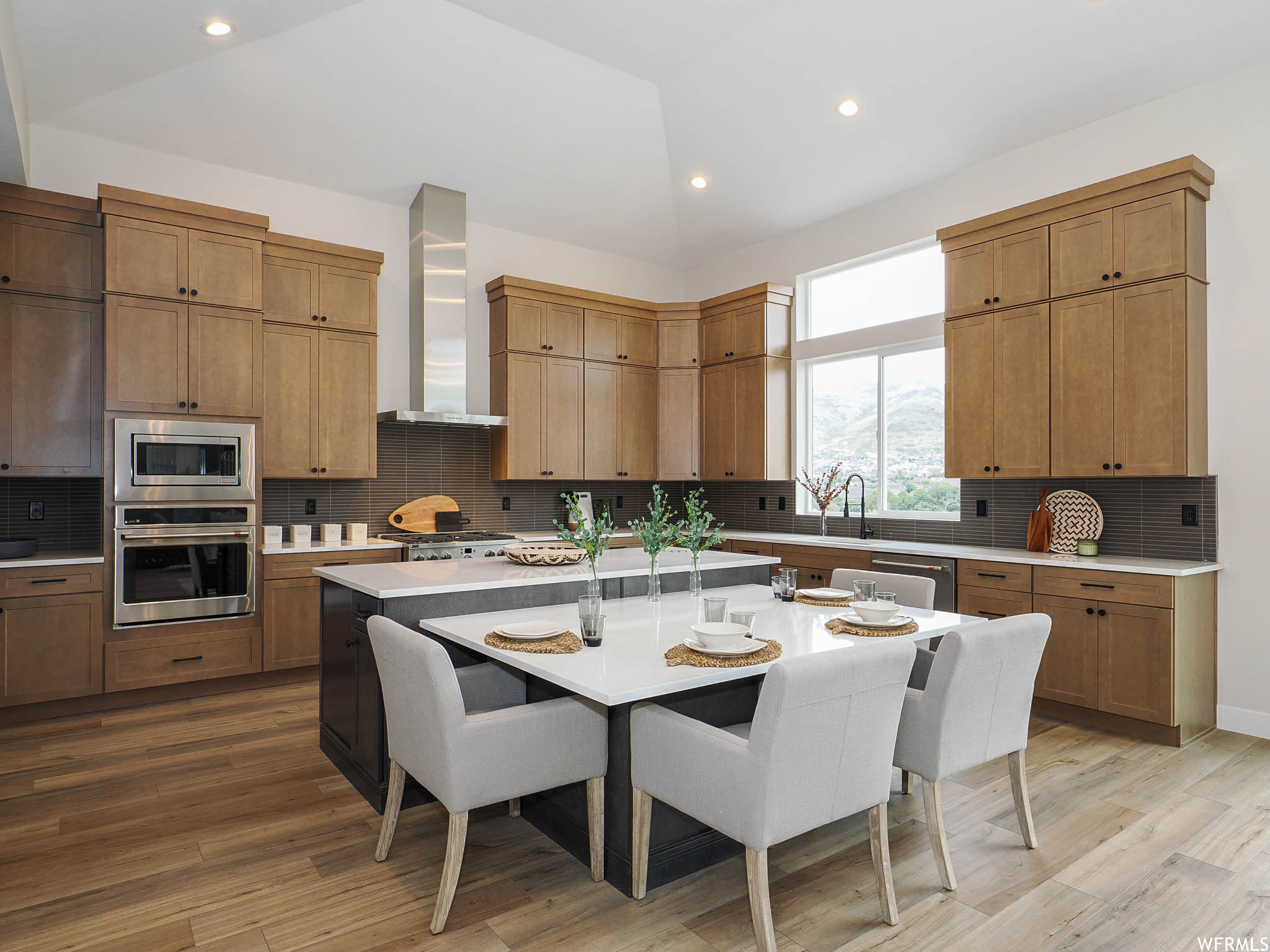 Kitchen with appliances with stainless steel finishes, wall chimney exhaust hood, light countertops, vaulted ceiling, light hardwood flooring, a center island with sink, and backsplash