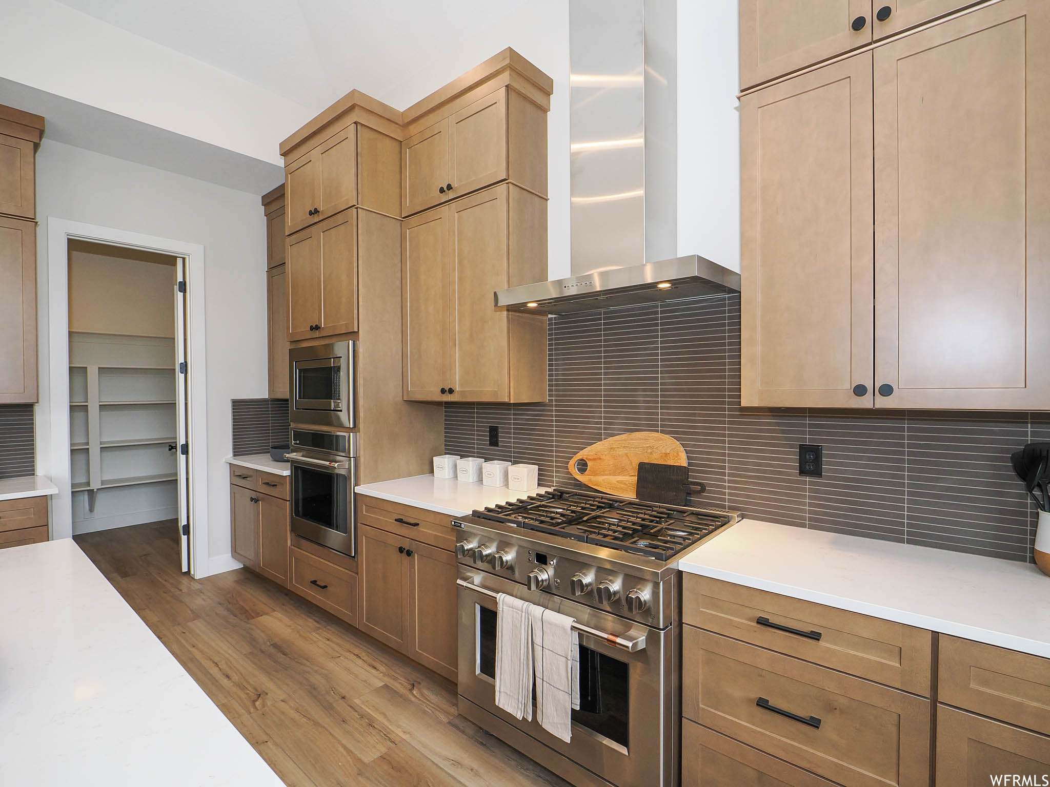 Kitchen featuring appliances with stainless steel finishes, wall chimney range hood, brown cabinets, light countertops, backsplash, and light hardwood floors