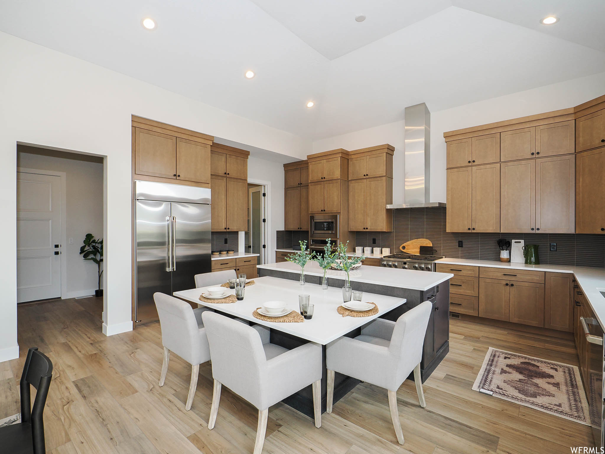 Kitchen featuring wall chimney exhaust hood, built in appliances, brown cabinets, light countertops, light hardwood flooring, and backsplash