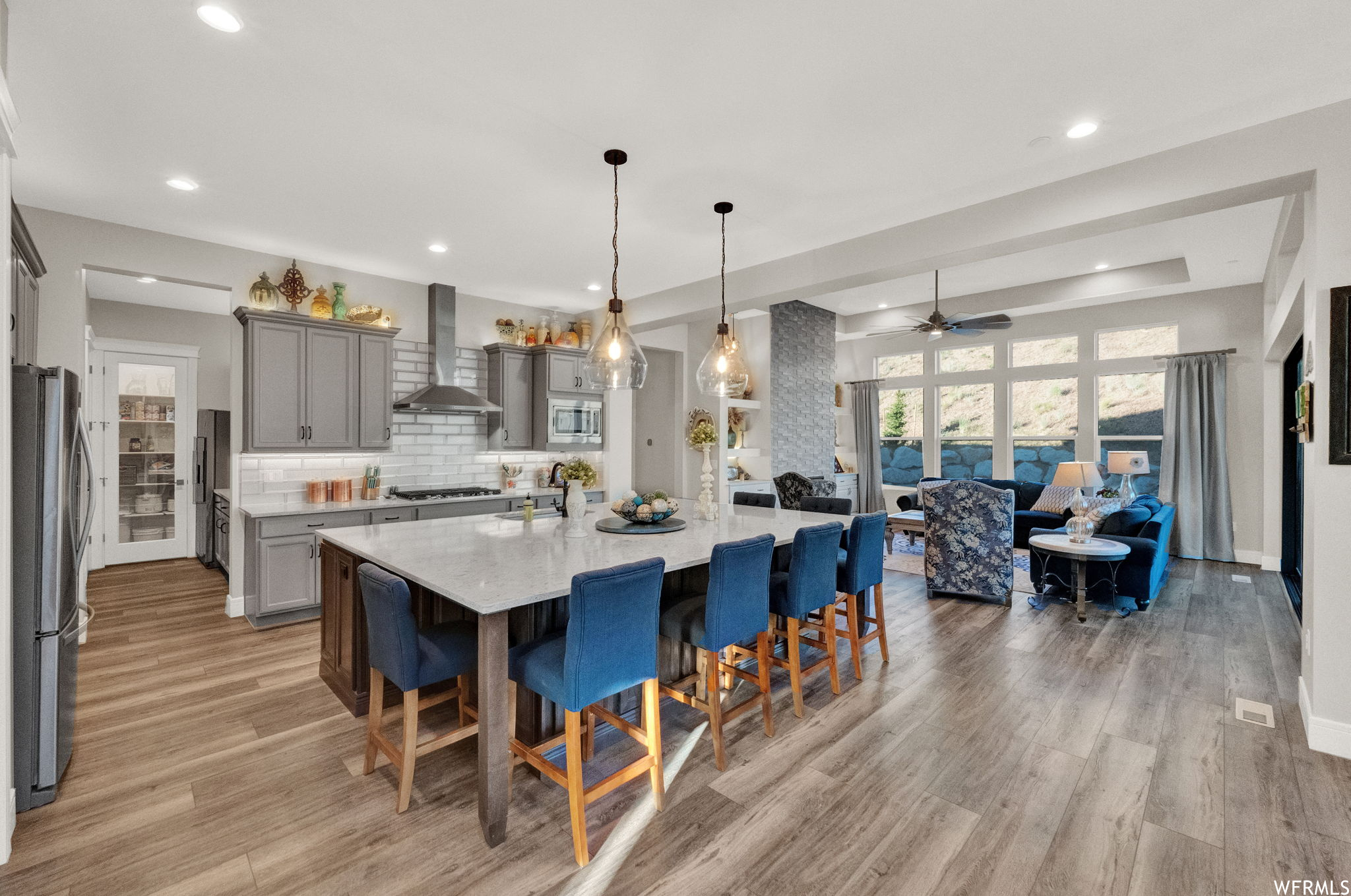 Kitchen with light hardwood flooring, decorative light fixtures, backsplash, ceiling fan, a center island, appliances with stainless steel finishes, a tray ceiling, kitchen island with sink, and wall chimney exhaust hood