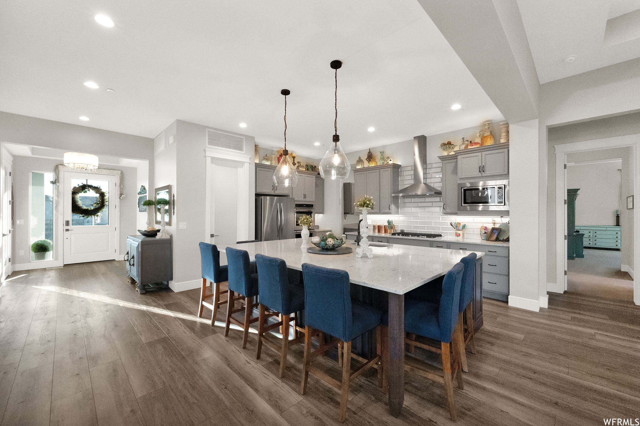 Kitchen featuring stainless steel appliances, backsplash, dark hardwood floors, a center island, pendant lighting, white cabinets, light countertops, kitchen island with sink, and wall chimney exhaust hood