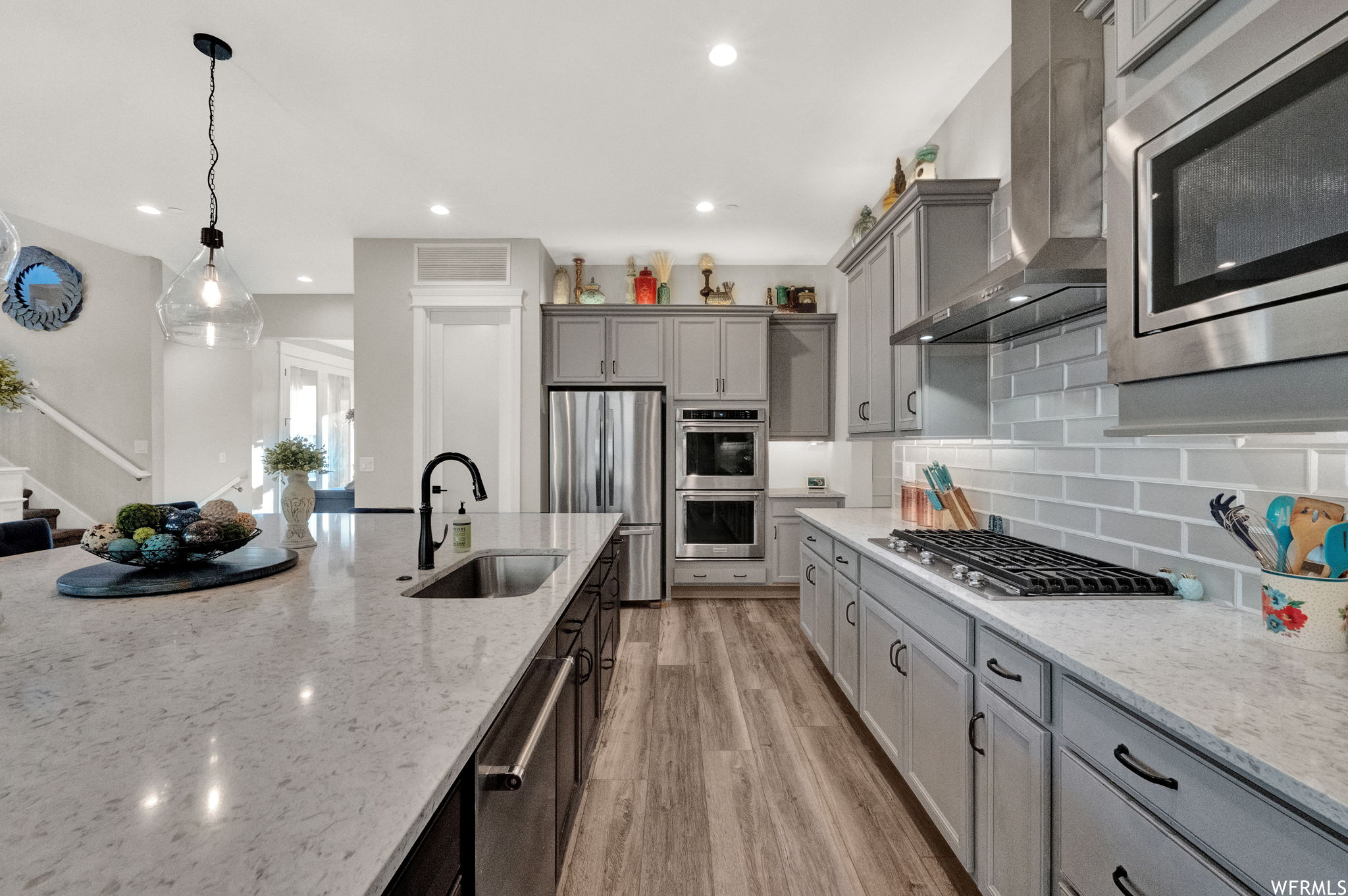 Kitchen with stainless steel appliances, wall chimney range hood, decorative light fixtures, wood-type flooring, white cabinets, backsplash, and light granite-like countertops