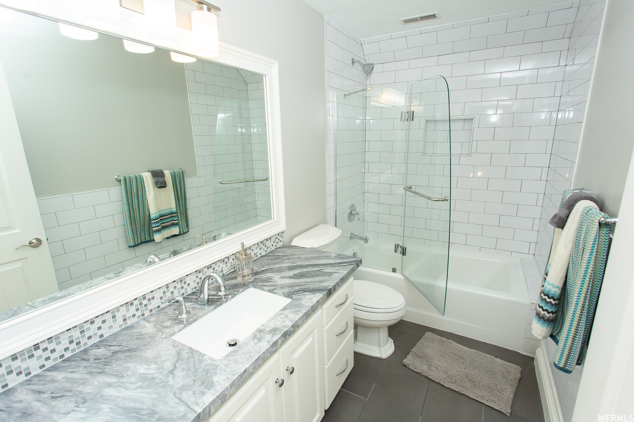 Full bathroom with light tile flooring, shower / bath combination with glass door, mirror, and vanity with extensive cabinet space.