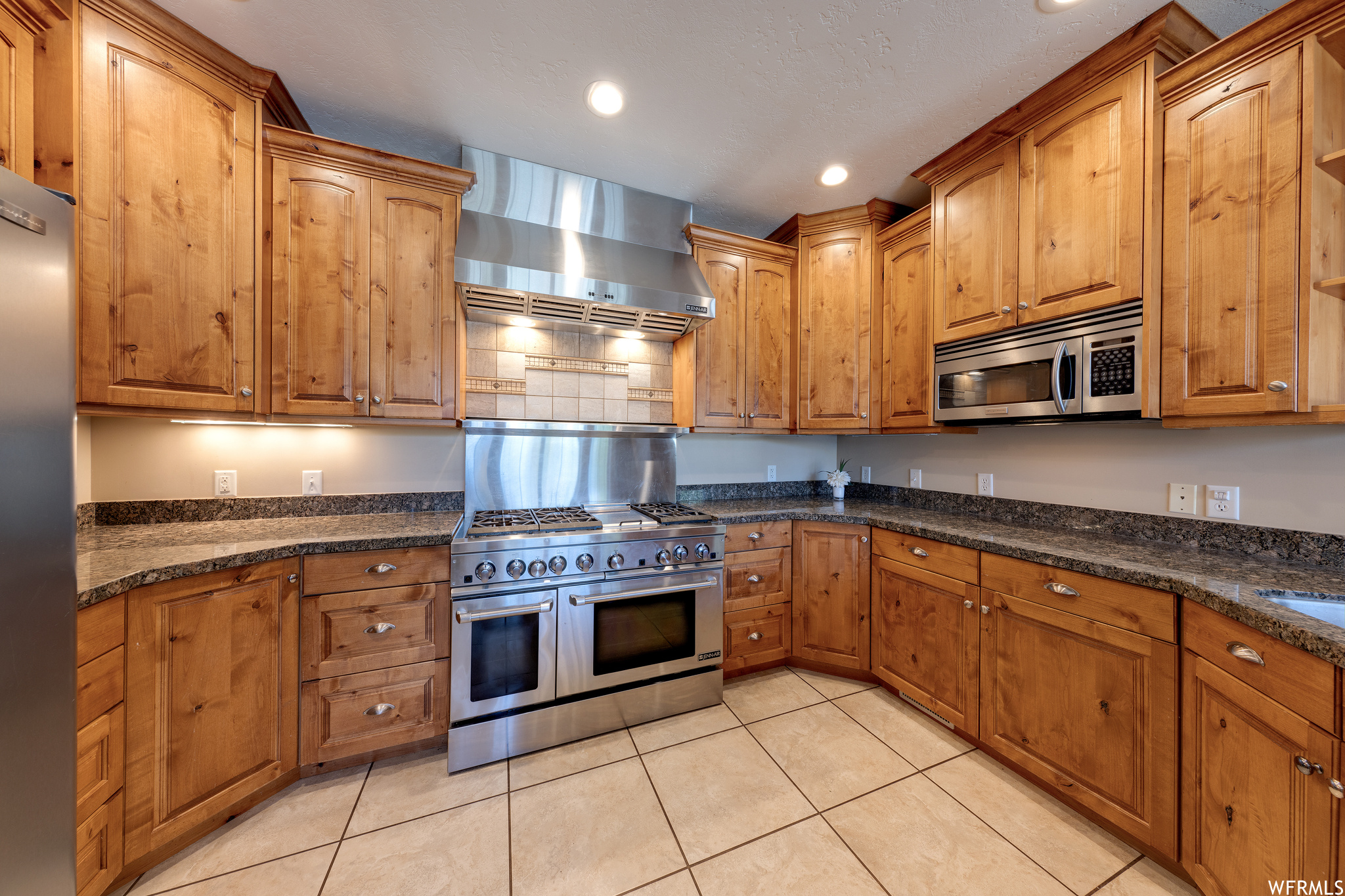 Kitchen featuring appliances with stainless steel finishes, wall chimney range hood, dark stone countertops, brown cabinets, and light tile floors