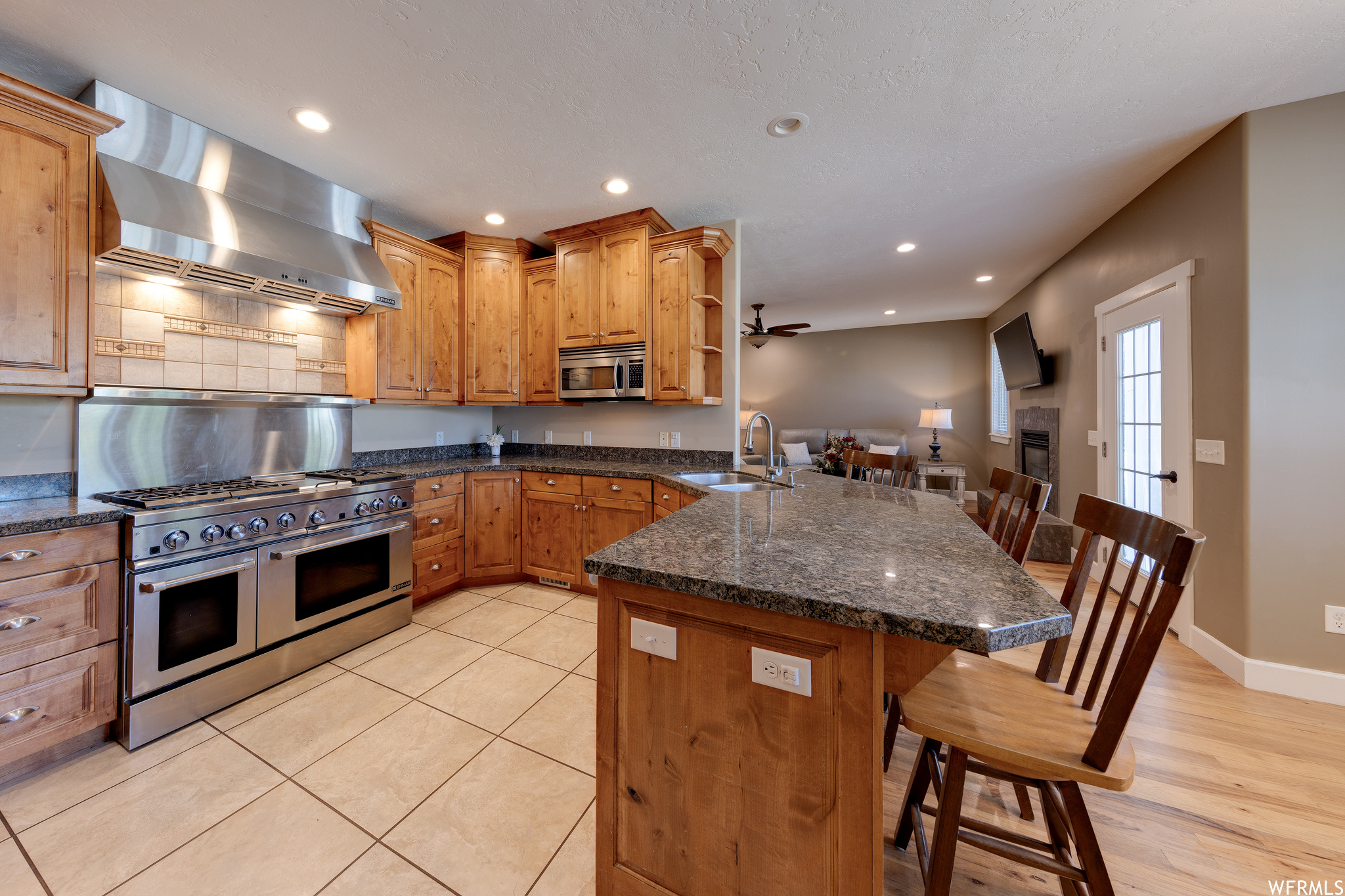 Kitchen featuring backsplash, brown cabinets, wall chimney range hood, light tile floors, appliances with stainless steel finishes, dark stone countertops, a center island, and ceiling fan