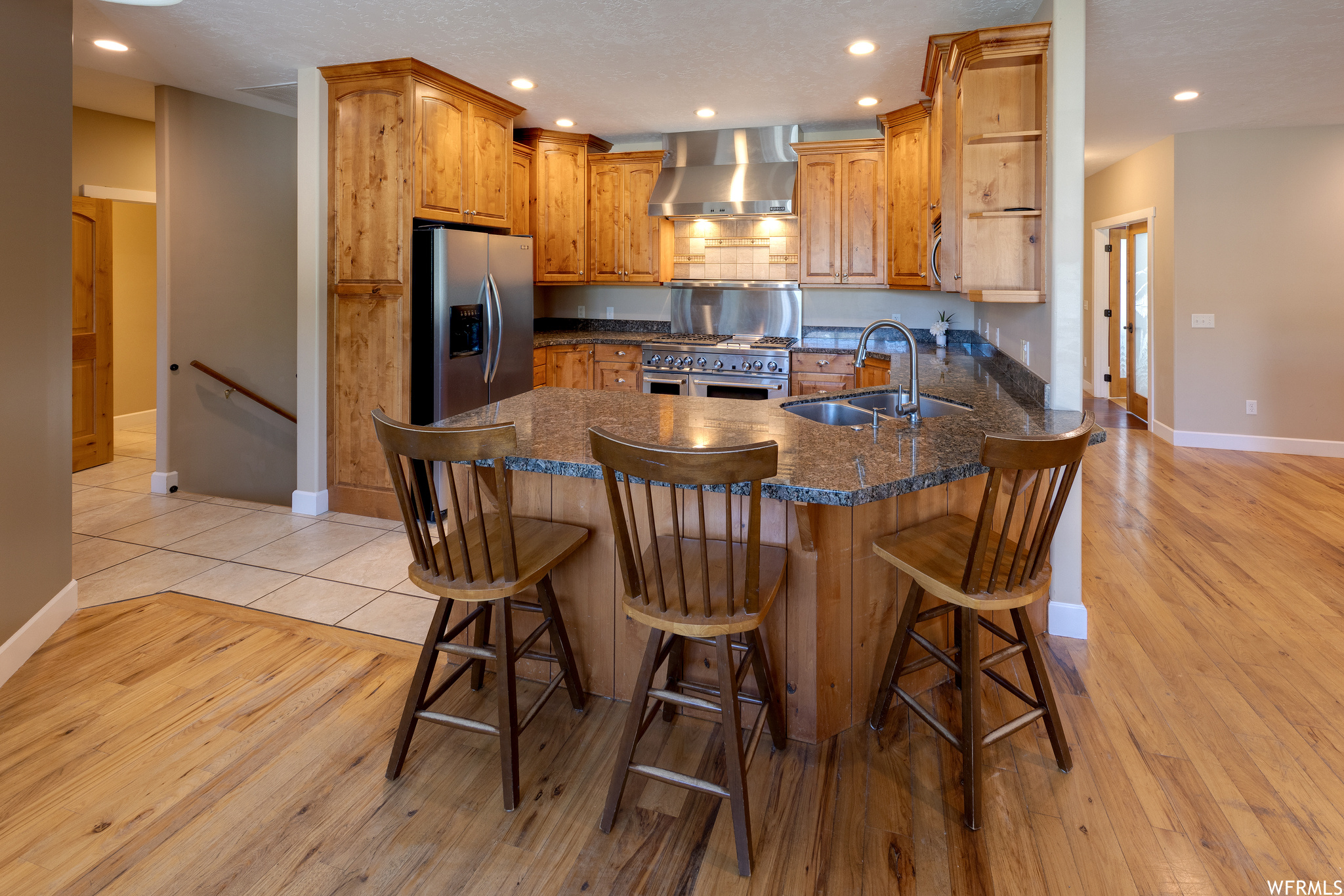 Kitchen with brown cabinets, wall chimney range hood, light hardwood flooring, a center island, stainless steel appliances, and dark stone counters