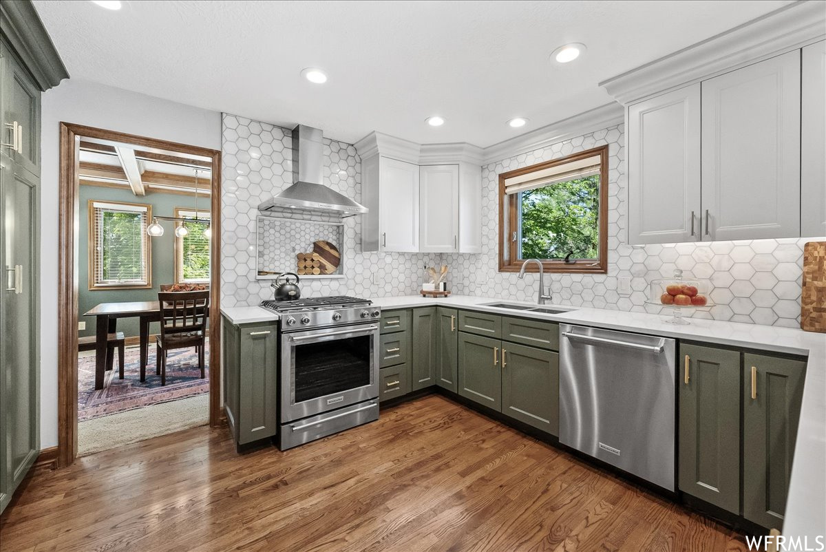 Kitchen featuring appliances with stainless steel finishes, plenty of natural light, wall chimney exhaust hood, light stone countertops, light hardwood flooring, and backsplash