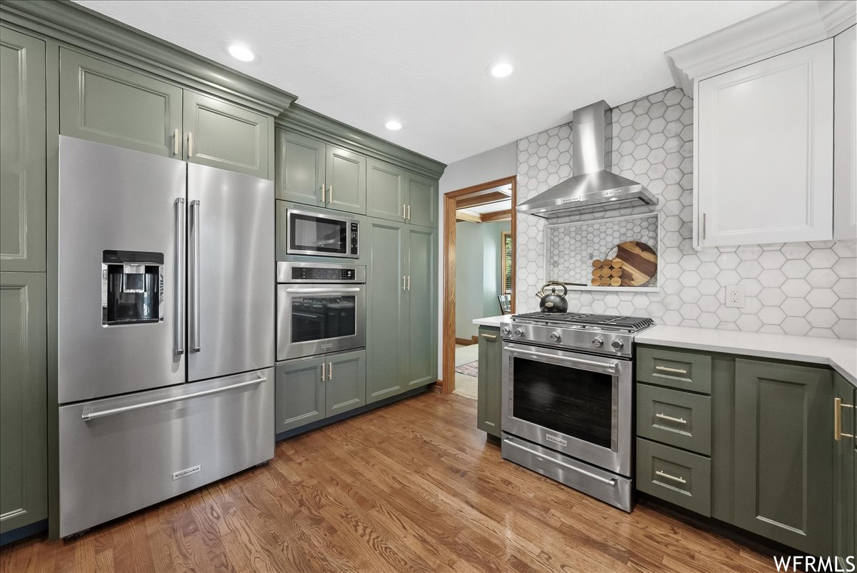 Kitchen featuring stainless steel appliances, wall chimney exhaust hood, light countertops, backsplash, and light hardwood floors