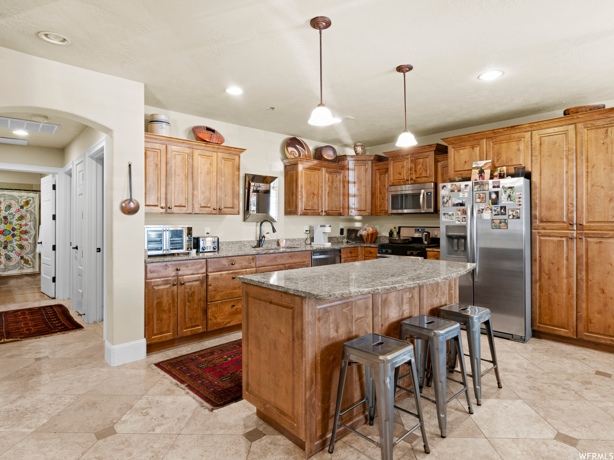 Kitchen with appliances with stainless steel finishes, decorative light fixtures, a center island, light stone counters, wood cabinets, and light tile floors