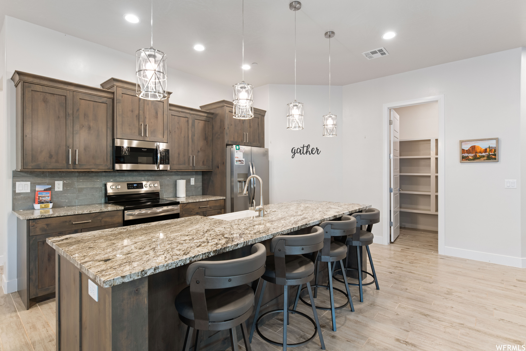 Kitchen featuring light hardwood / wood-style floors, an island with sink, backsplash, decorative light fixtures, and appliances with stainless steel finishes