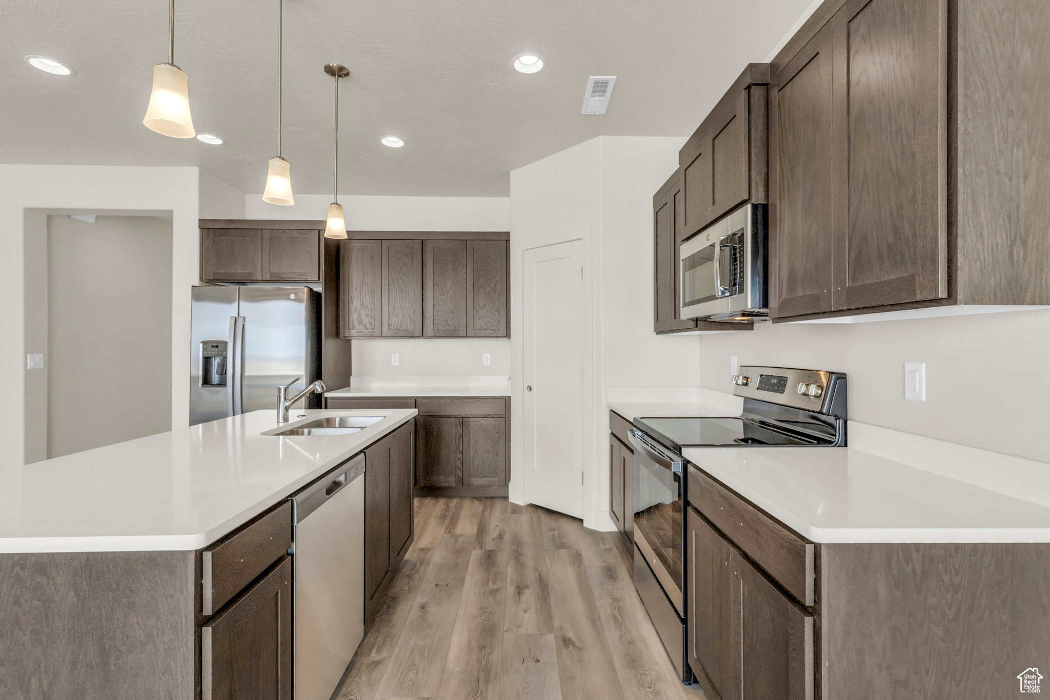 Kitchen featuring light wood-type flooring, pendant lighting, dark brown cabinets, appliances with stainless steel finishes, and an island with sink