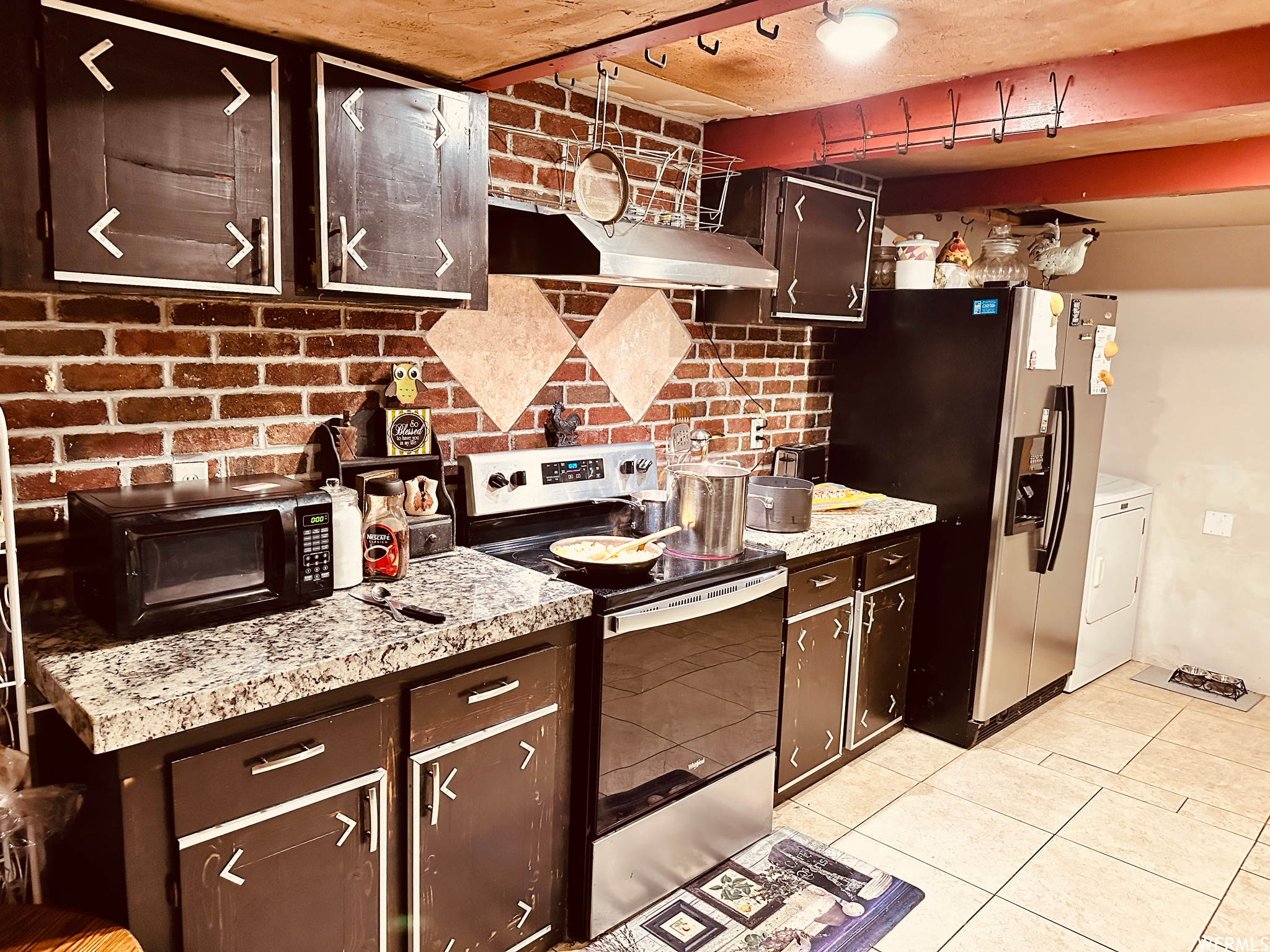 Kitchen with stainless steel appliances, dark brown cabinets, light tile floors, and brick wall
