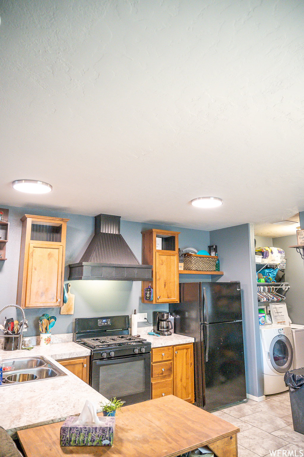 Kitchen with washer / clothes dryer, sink, wall chimney range hood, light tile floors, and black appliances