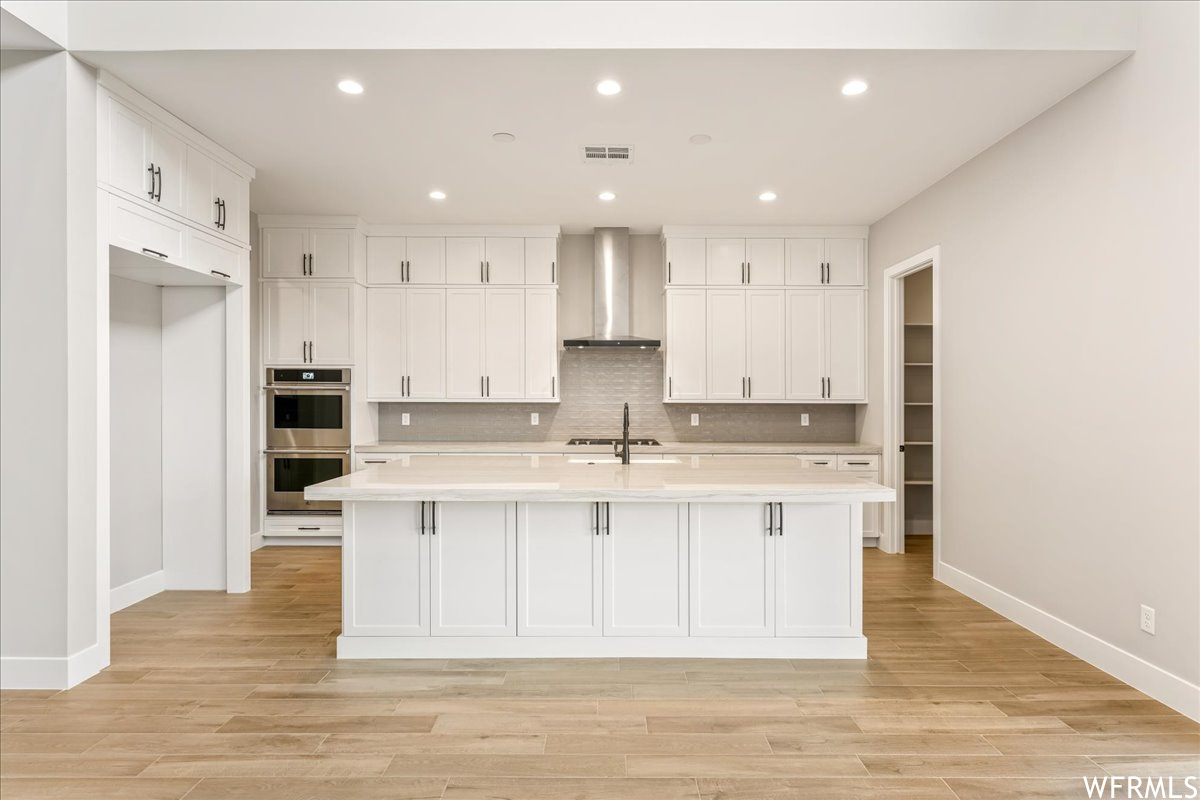 Kitchen featuring wall chimney range hood, a kitchen island with sink, light hardwood / wood-style floors, stainless steel double oven, and white cabinetry