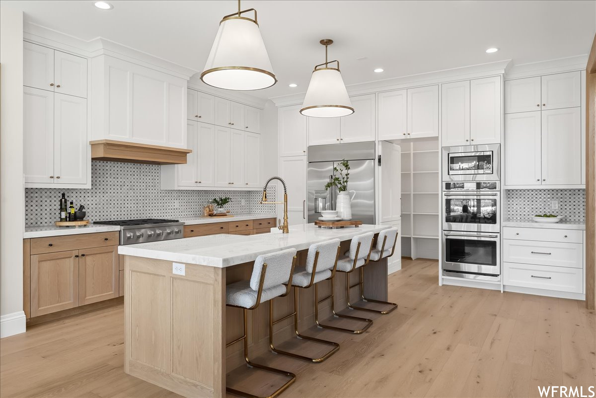 Kitchen featuring light hardwood / wood-style flooring, a kitchen island with sink, built in appliances, hanging light fixtures, and backsplash