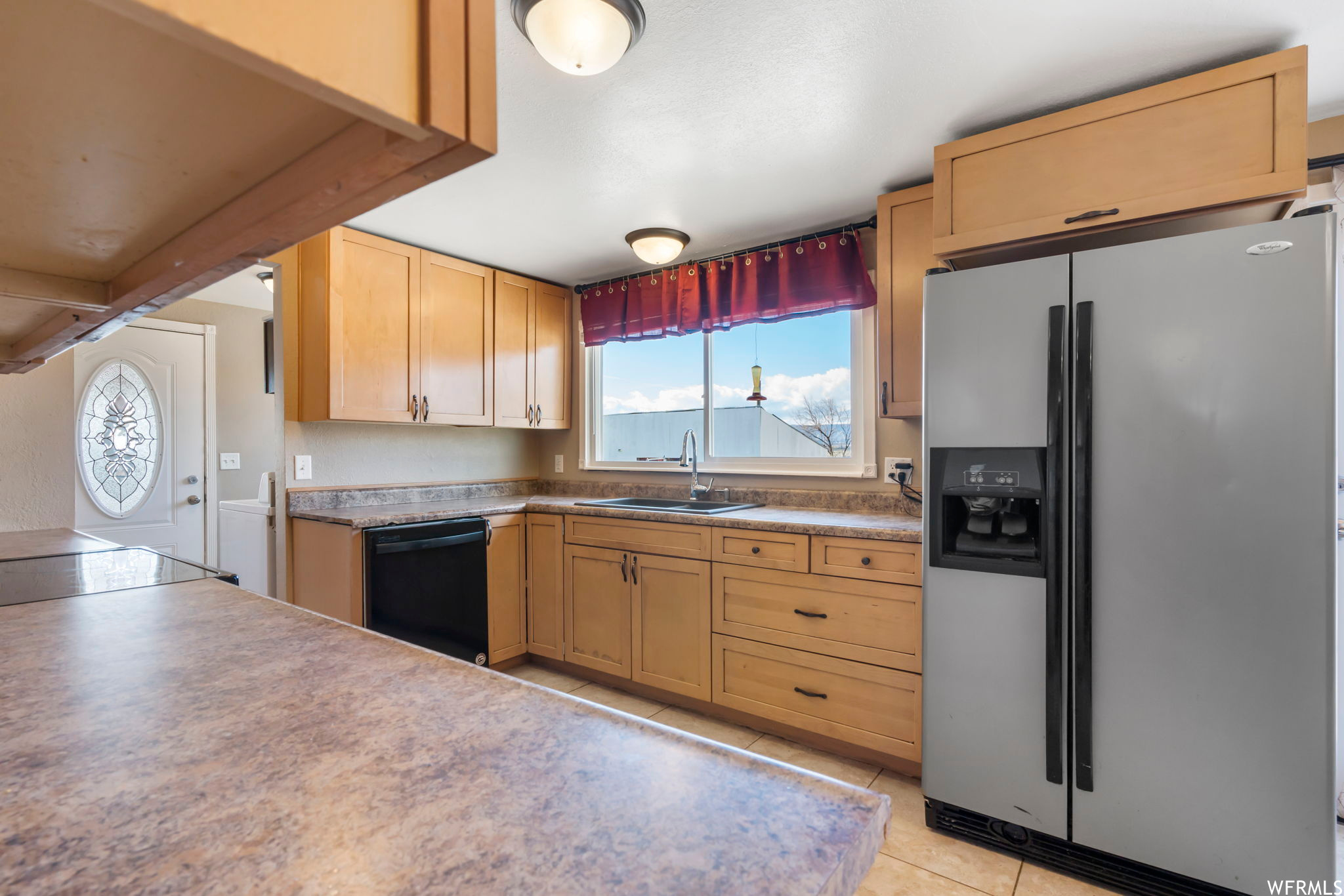 Kitchen featuring light tile flooring, sink, stainless steel fridge with ice dispenser, black dishwasher, and light brown cabinetry