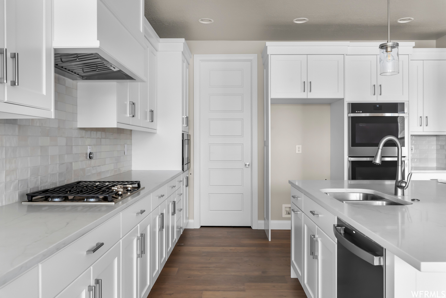 Kitchen featuring dark wood-type flooring, custom exhaust hood, stainless steel appliances, decorative light fixtures, and backsplash