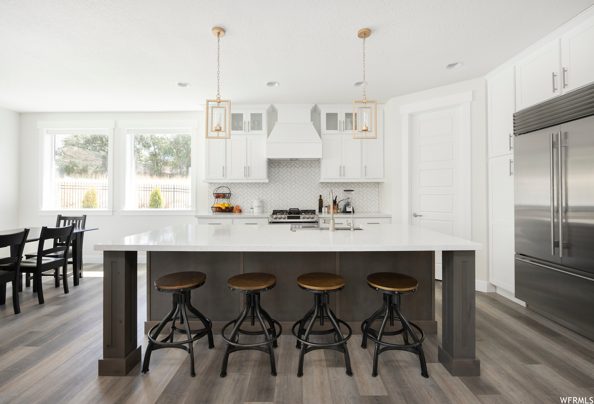 Kitchen with hanging light fixtures, custom range hood, a kitchen island with sink, and appliances with stainless steel finishes
