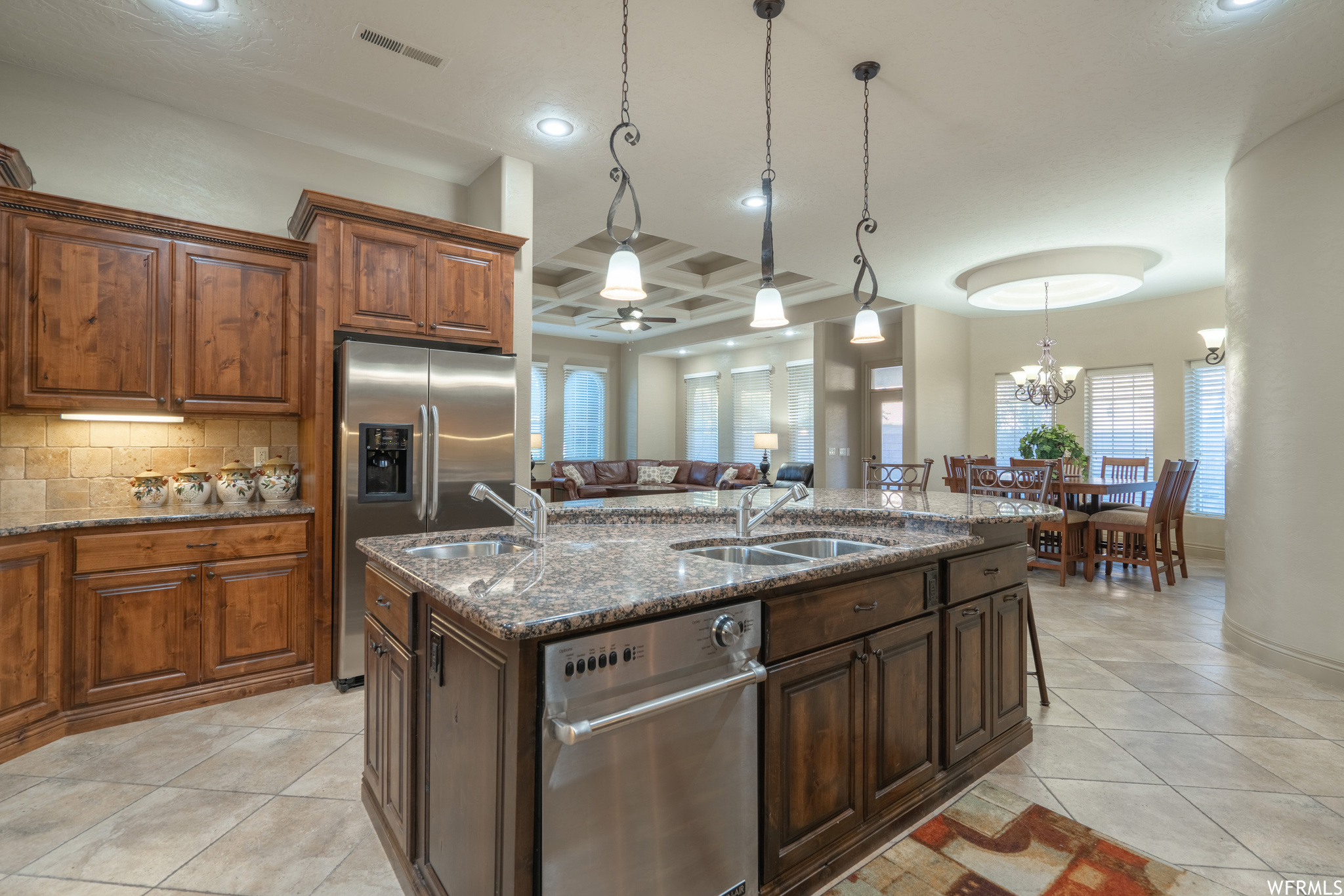 Kitchen featuring stainless steel dishwasher, hanging light fixtures, coffered ceiling, tasteful backsplash, and an island with sink