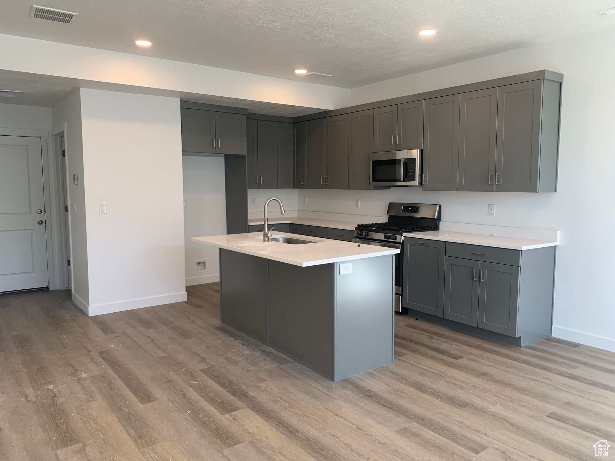 Kitchen with gray cabinets, an island with sink, light hardwood / wood-style floors, and appliances with stainless steel finishes