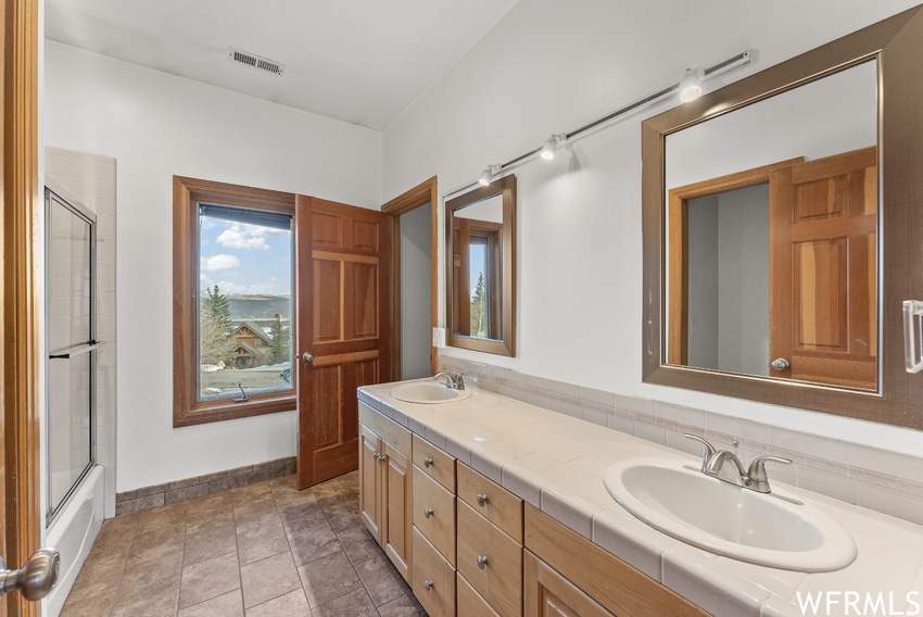Bathroom featuring tile floors, double sink vanity, tasteful backsplash, and shower / bath combination with glass door