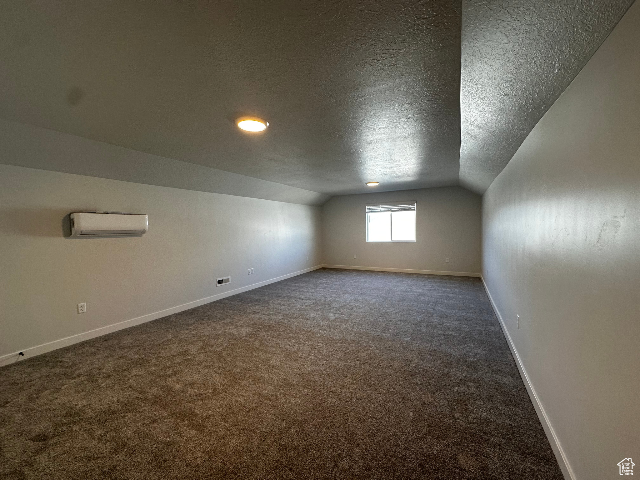 Additional living space with a textured ceiling, vaulted ceiling, an AC wall unit, and dark colored carpet