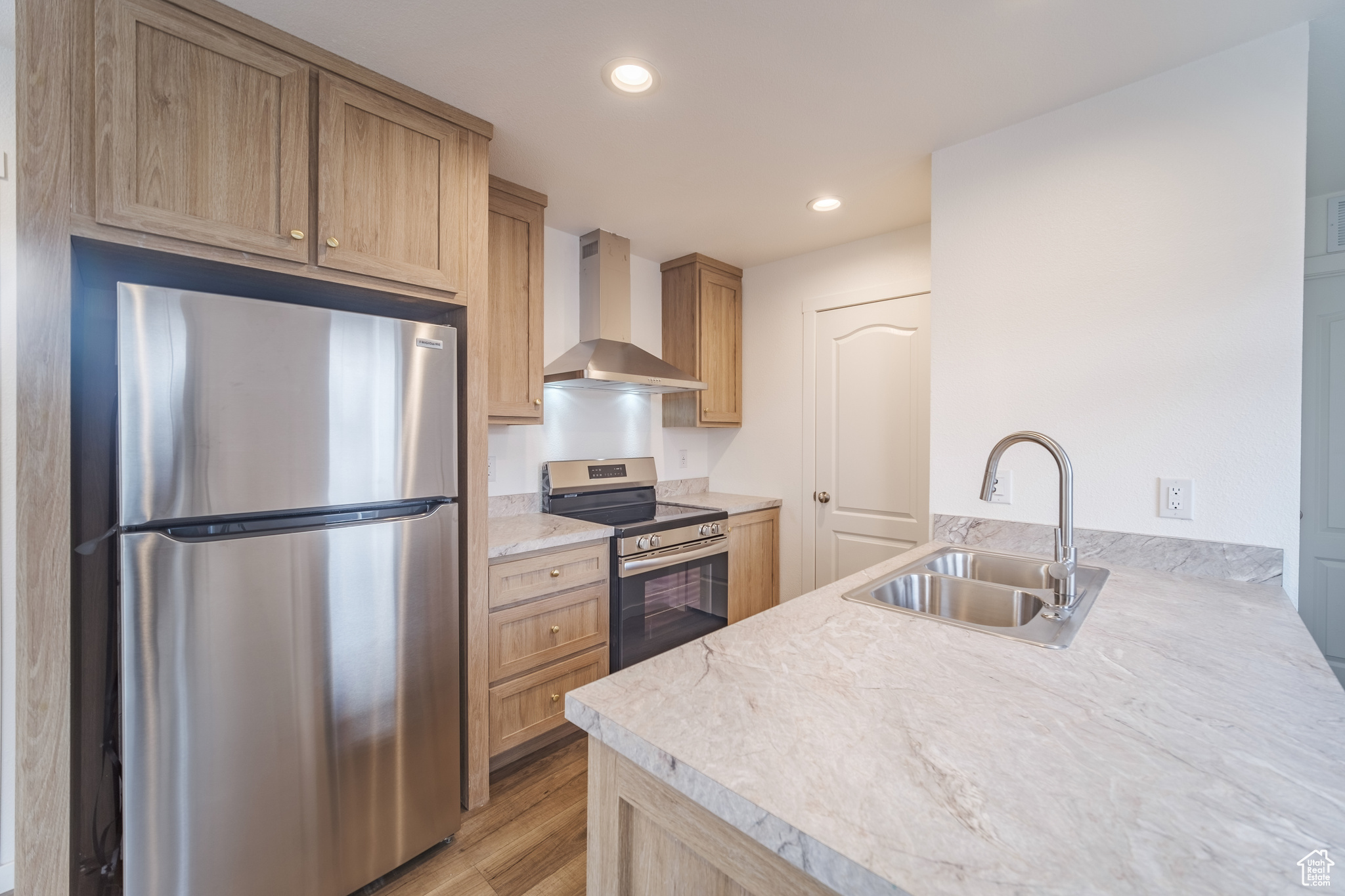 Kitchen with wall chimney exhaust hood, appliances with stainless steel finishes, light brown cabinetry, sink, and light wood-type flooring