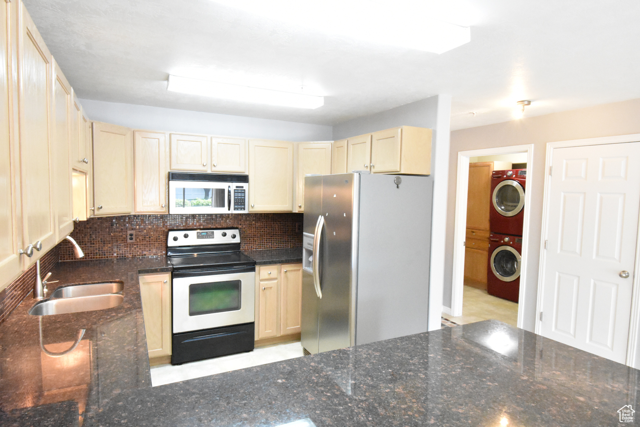 Kitchen with dark stone countertops, stainless steel appliances, stacked washer and clothes dryer, sink, and tasteful backsplash