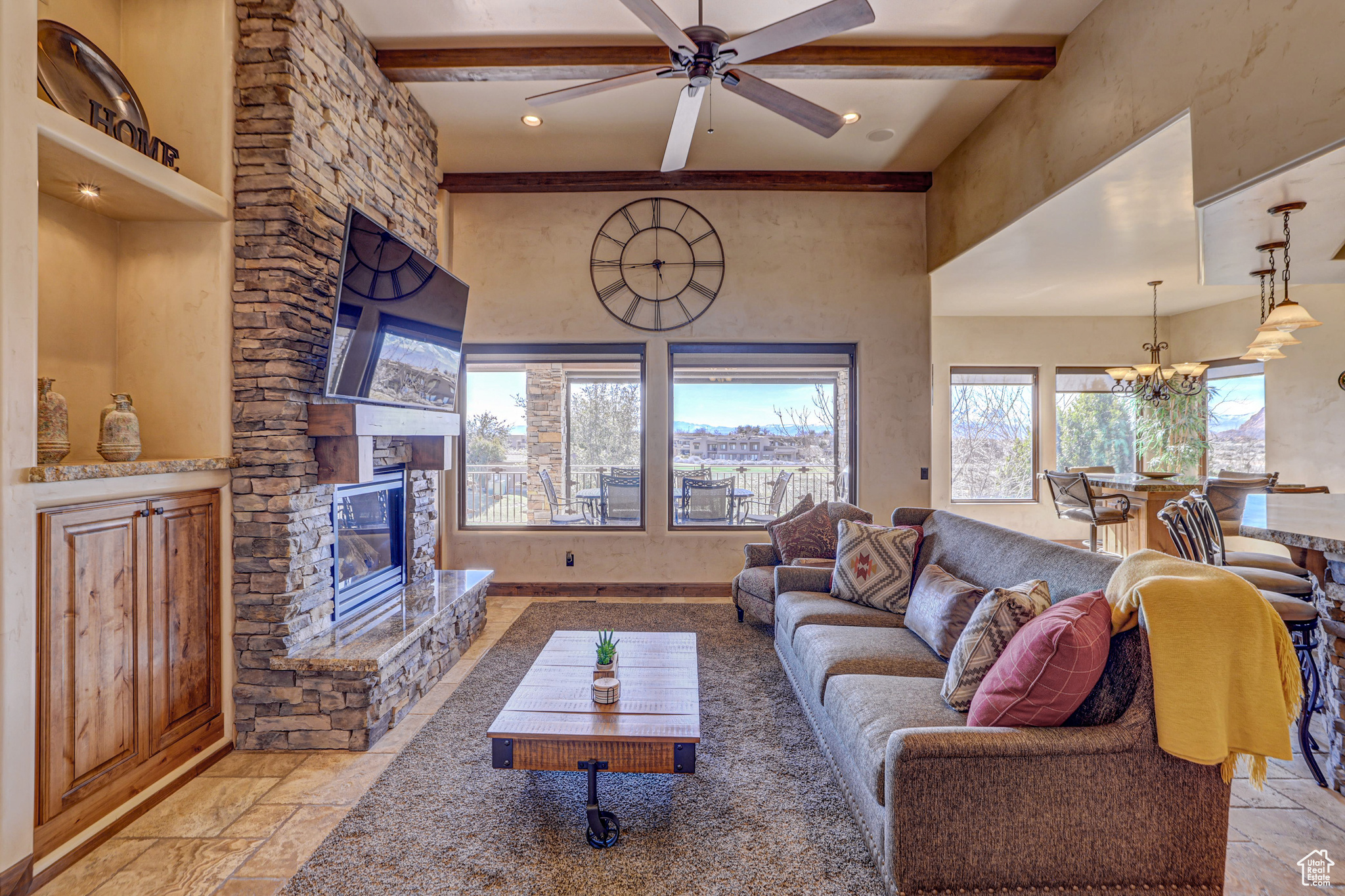 Living room featuring a stone fireplace, beamed ceiling, ceiling fan with notable chandelier, and light tile floors