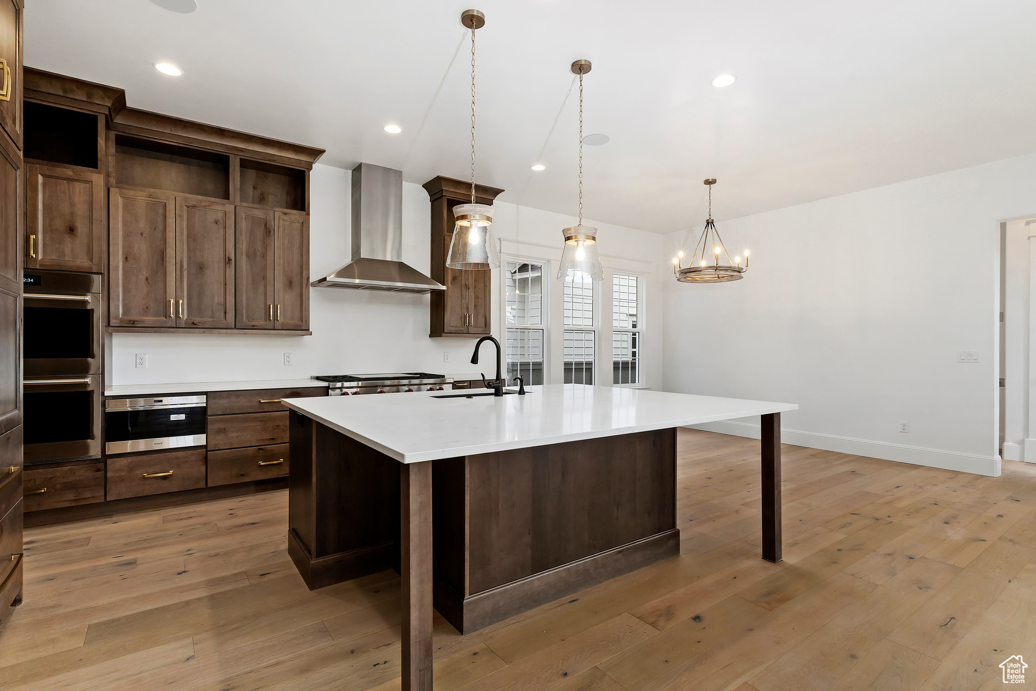 Kitchen with light wood-type flooring, pendant lighting, a notable chandelier, appliances with stainless steel finishes, and wall chimney range hood
