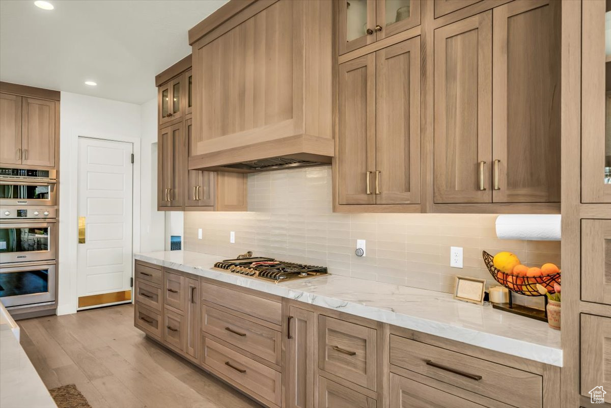 Kitchen featuring stainless steel appliances, light stone counters, custom range hood, tasteful backsplash, and light hardwood / wood-style floors