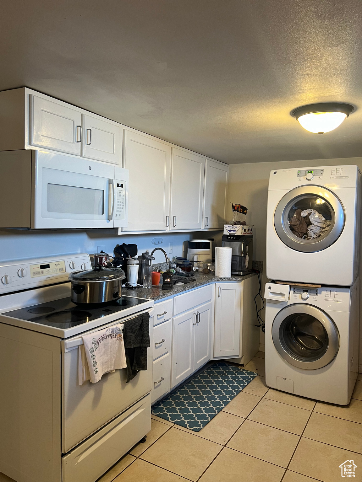 Kitchen with white cabinets, light tile floors, white appliances, and stacked washer and clothes dryer