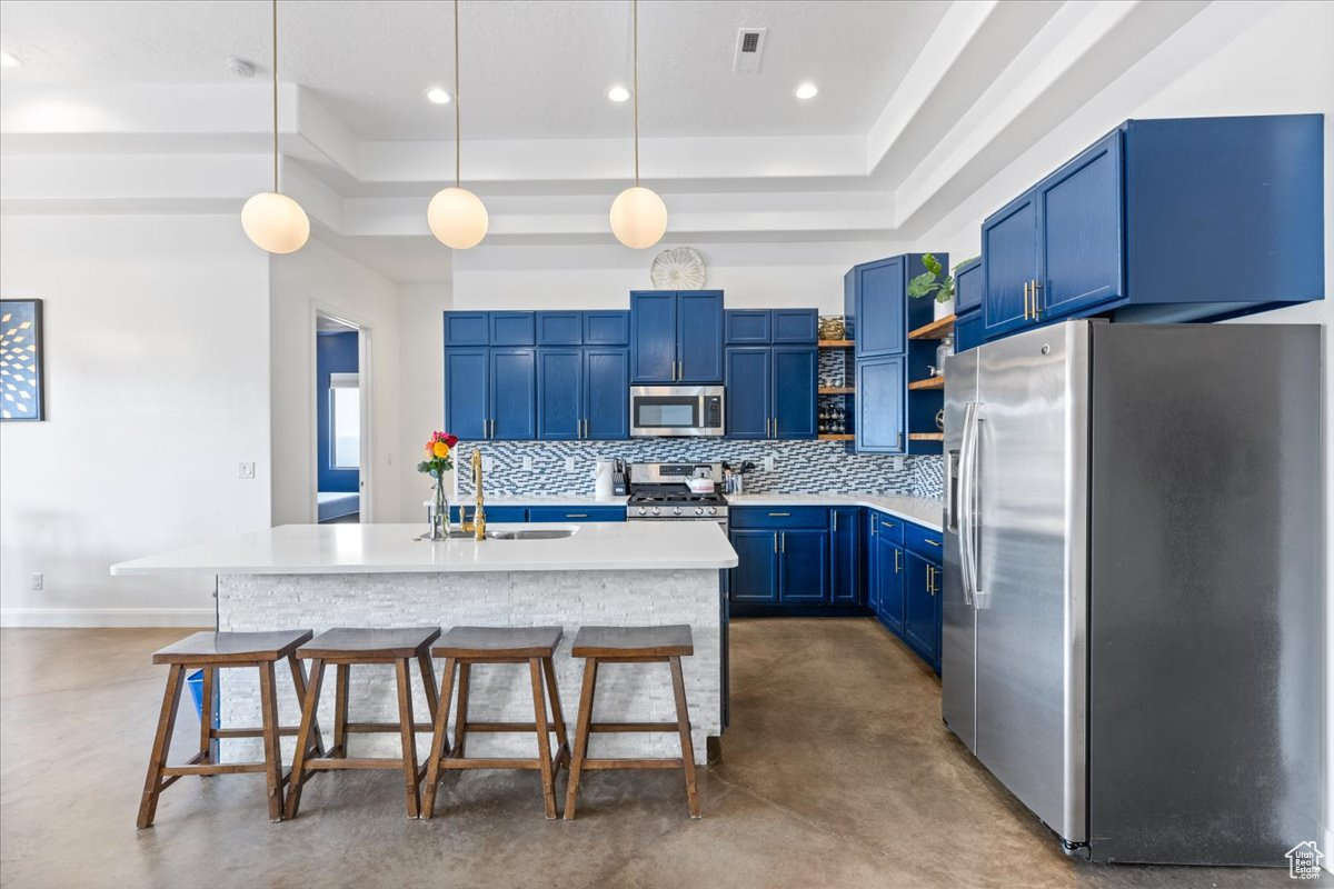 Kitchen with tasteful backsplash, a tray ceiling, an island with sink, hanging light fixtures, and stainless steel appliances