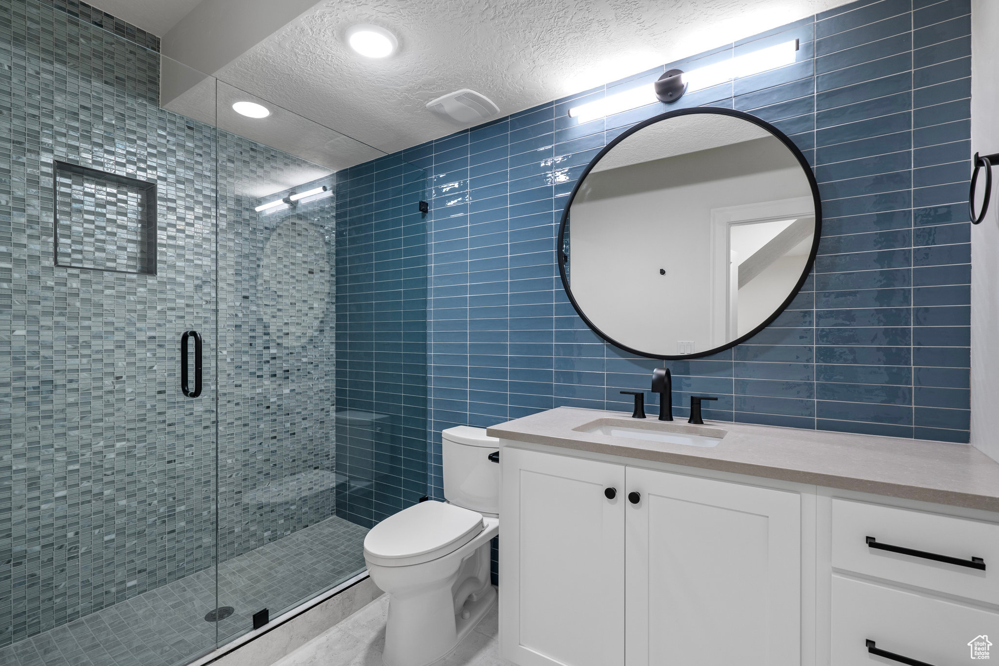 Bathroom featuring a textured ceiling, tile walls, vanity with extensive cabinet space, a shower with door, and toilet