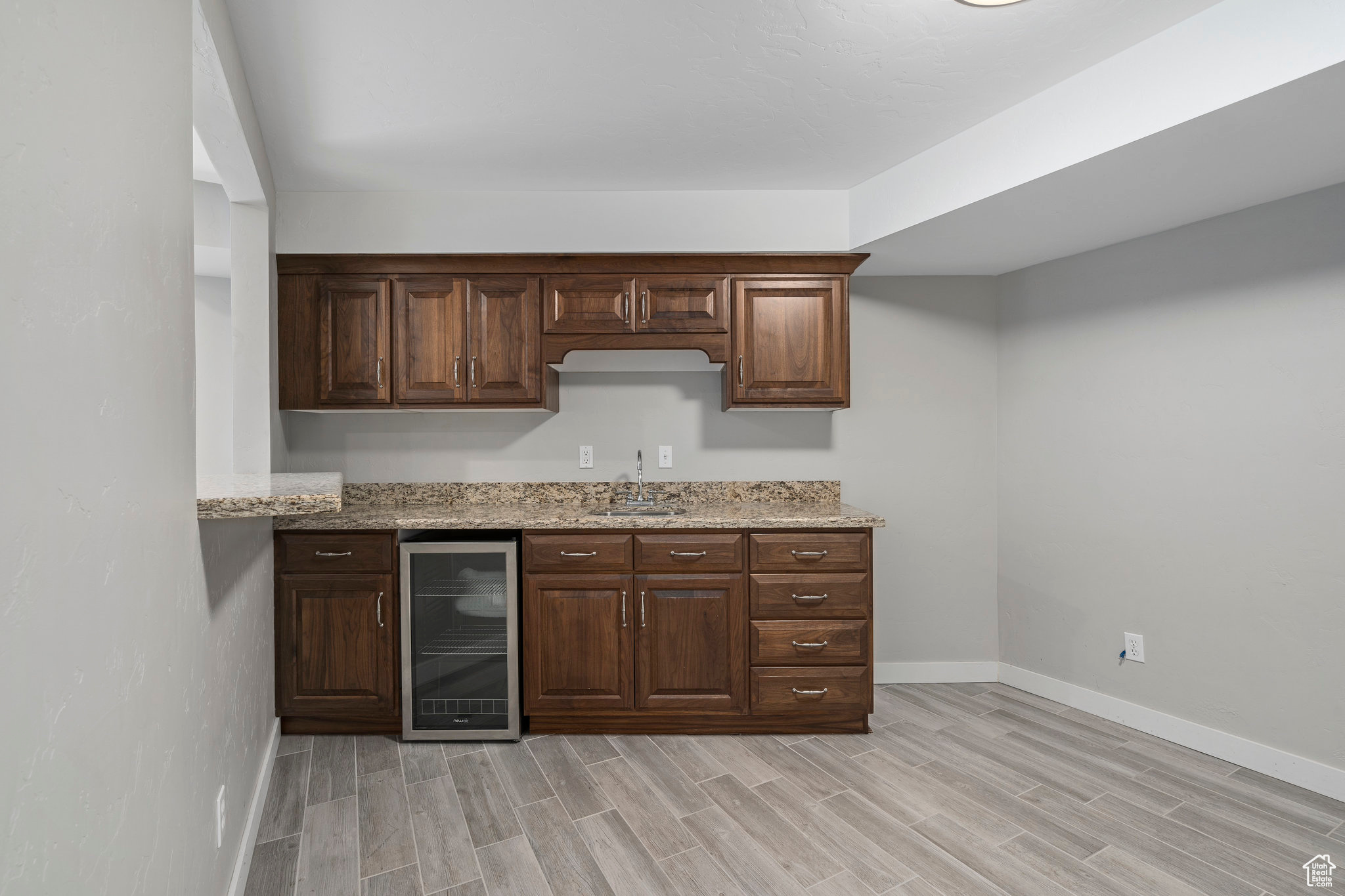 Basement kitchenette with murphy bed attached on adjacent wall. Room has light wood-style flooring, light granite countertops, beverage cooler, and dark brown cabinetry