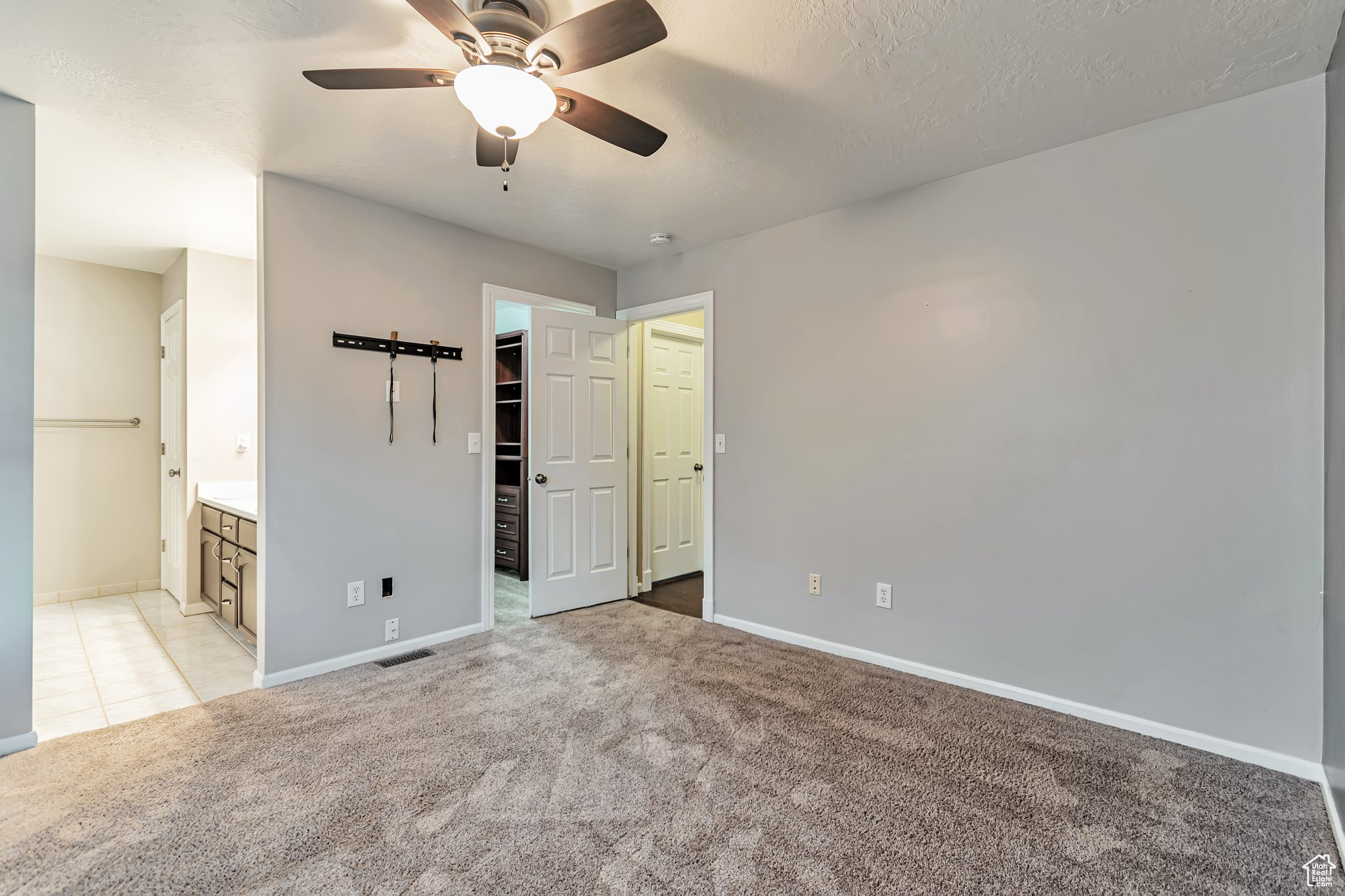 Master bedroom with a walk in closet with built in shelving, light carpet, ensuite bathroom, TV mount, and ceiling fan