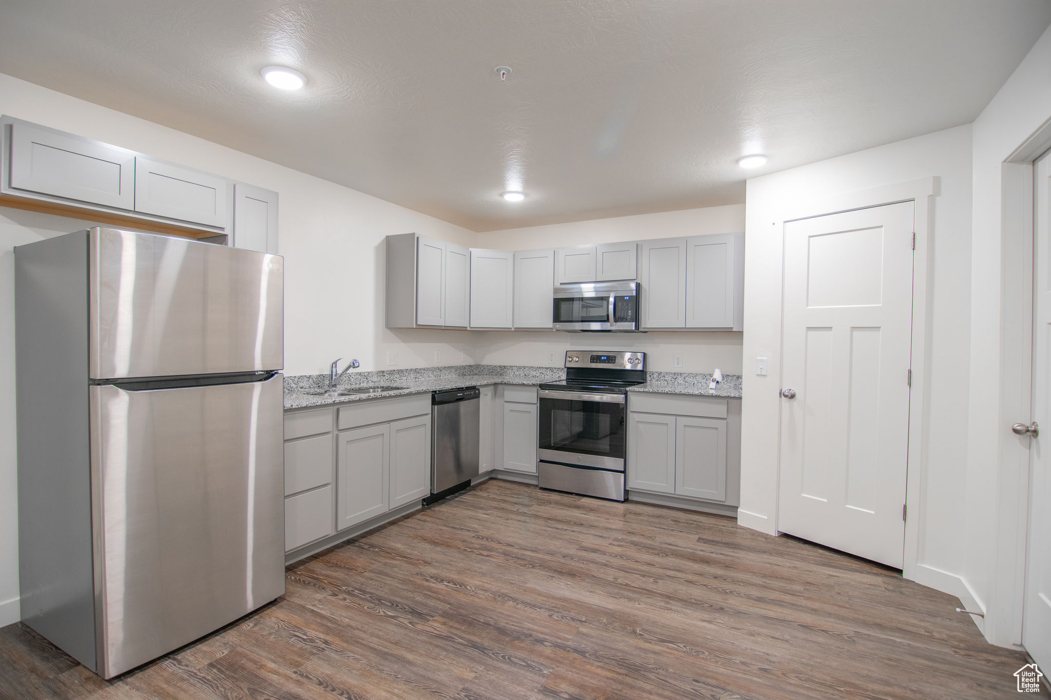 Kitchen with gray cabinets, appliances with stainless steel finishes, light stone counters, dark hardwood / wood-style floors, and sink