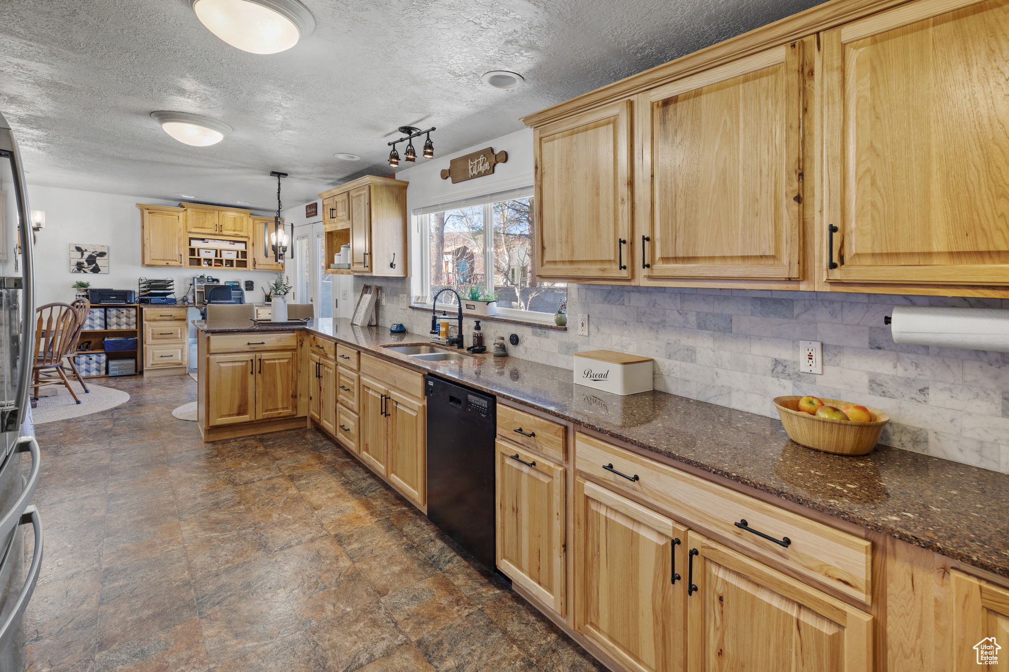 Kitchen with sink, dark stone counters, hanging light fixtures, black dishwasher, and a textured ceiling