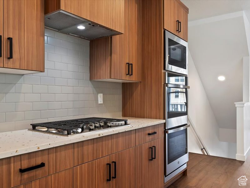 Kitchen featuring dark hardwood / wood-style floors, appliances with stainless steel finishes, light stone countertops, custom exhaust hood, and tasteful backsplash
