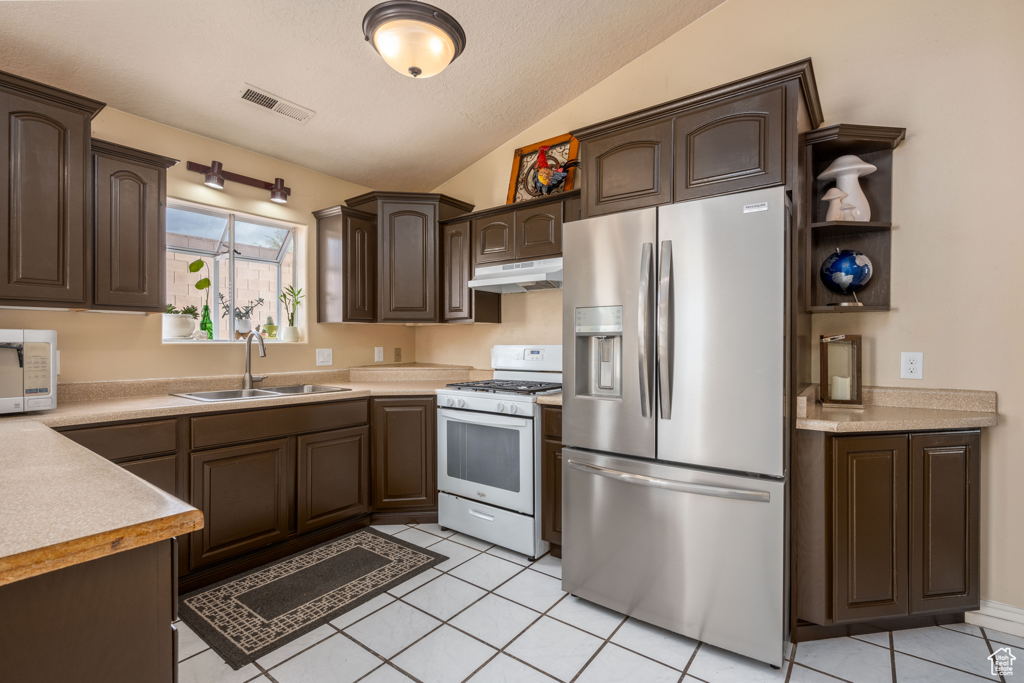 Kitchen featuring sink, white appliances, vaulted ceiling, dark brown cabinetry, and light tile floors