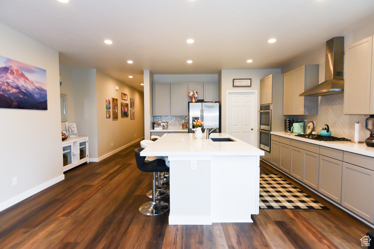 Kitchen featuring wall chimney range hood, tasteful backsplash, stainless steel appliances, dark hardwood / wood-style floors, and a kitchen island with sink
