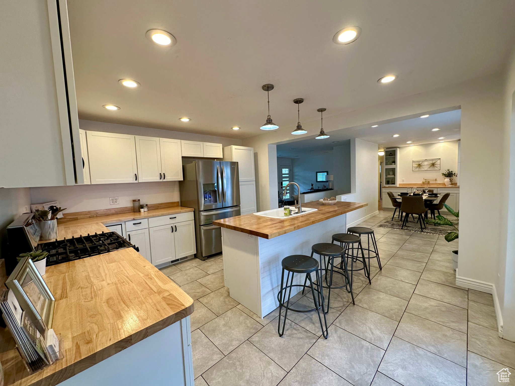 Kitchen featuring stainless steel fridge with ice dispenser, hanging light fixtures, an island with sink, white cabinets, and wood counters