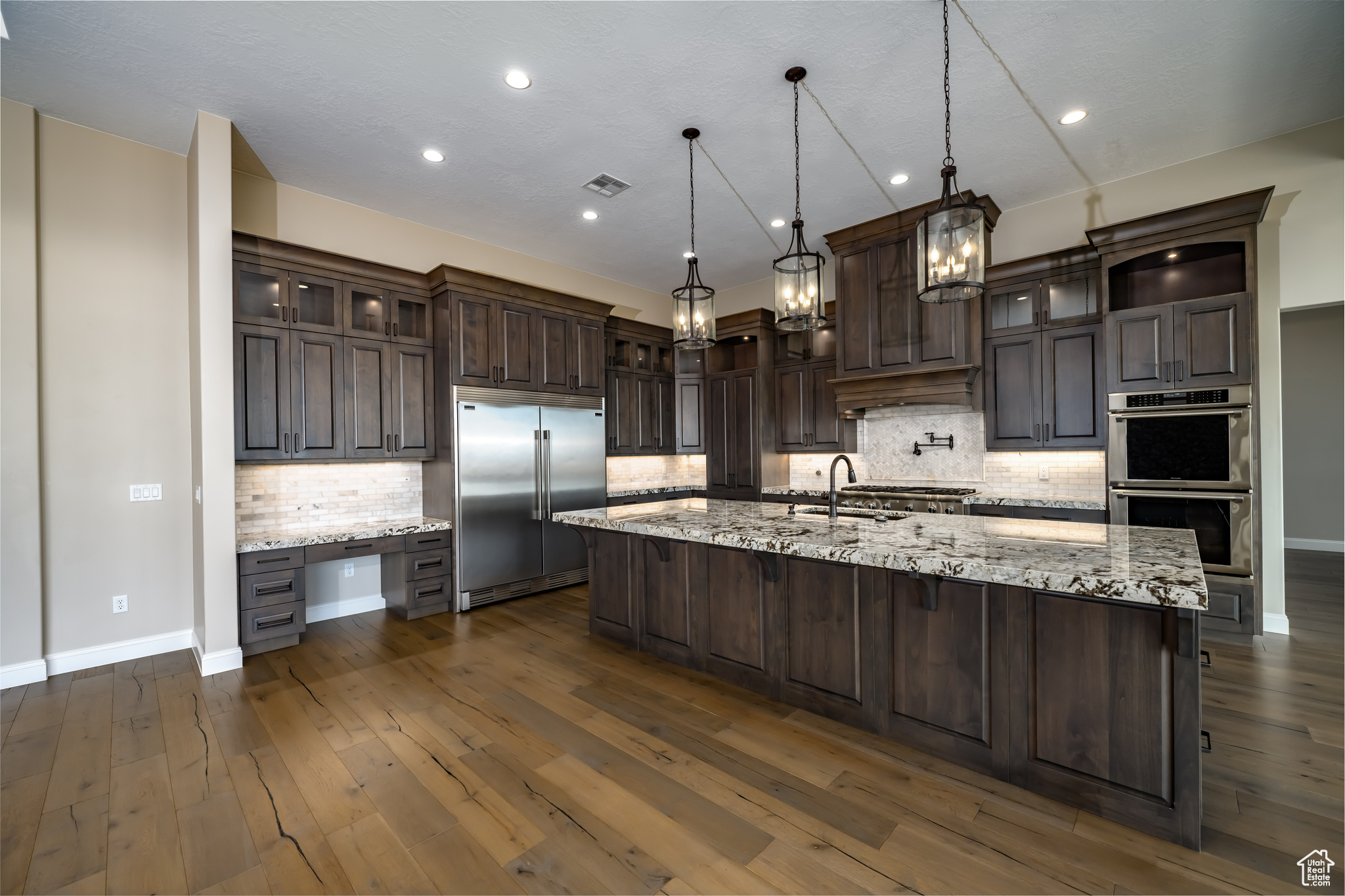 Kitchen with appliances with stainless steel finishes, backsplash, dark wood-type flooring, decorative light fixtures, and a kitchen island with sink