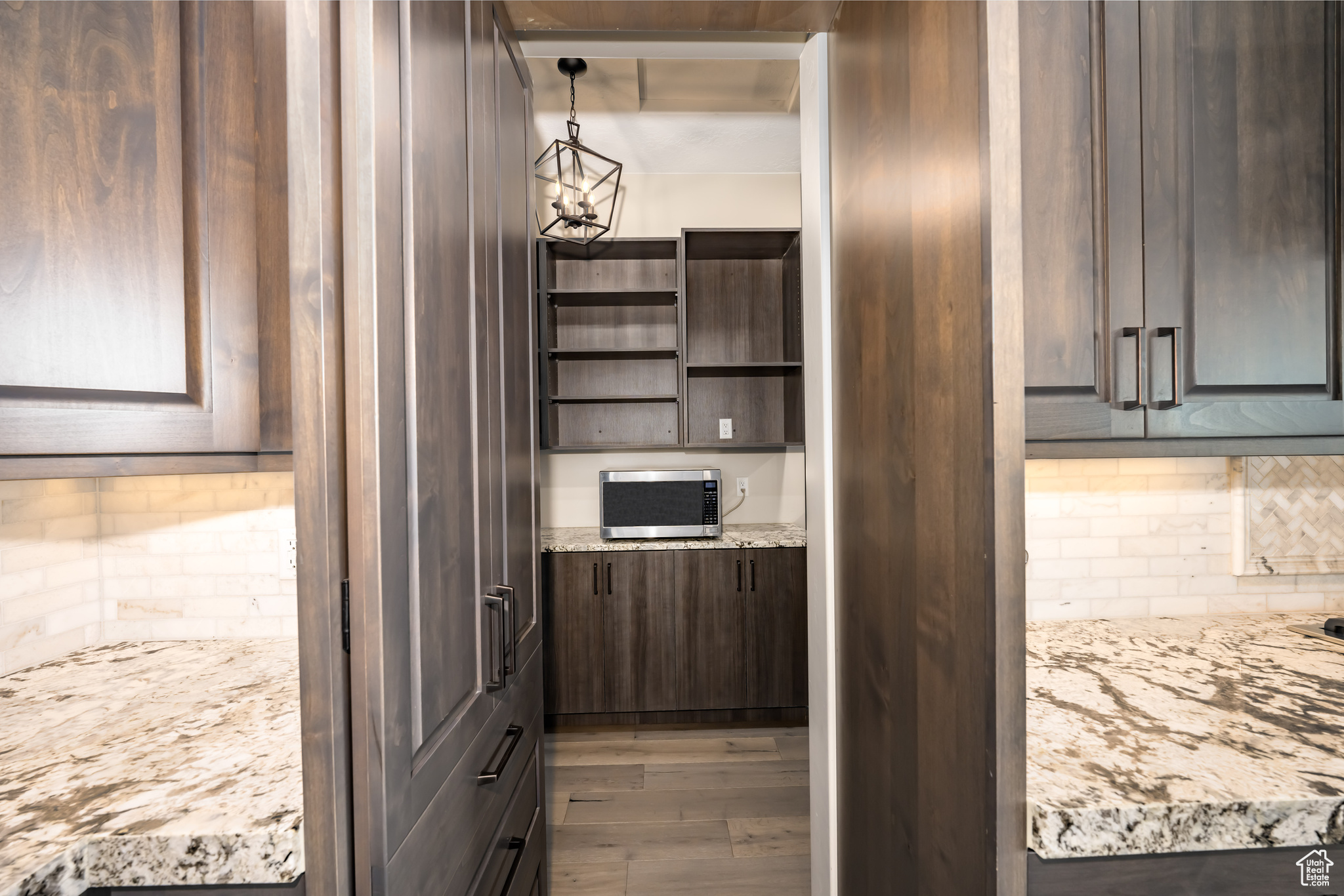 Kitchen with dark brown cabinets, backsplash, hardwood / wood-style flooring, light stone counters, and pendant lighting