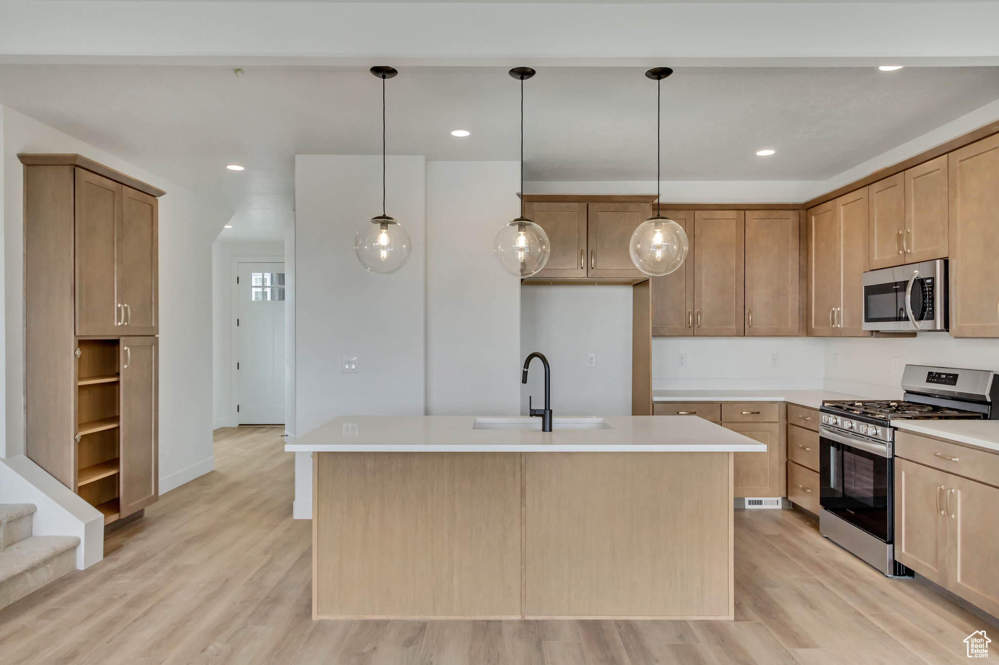 Kitchen featuring appliances with stainless steel finishes, sink, light wood-type flooring, hanging light fixtures, and a kitchen island with sink