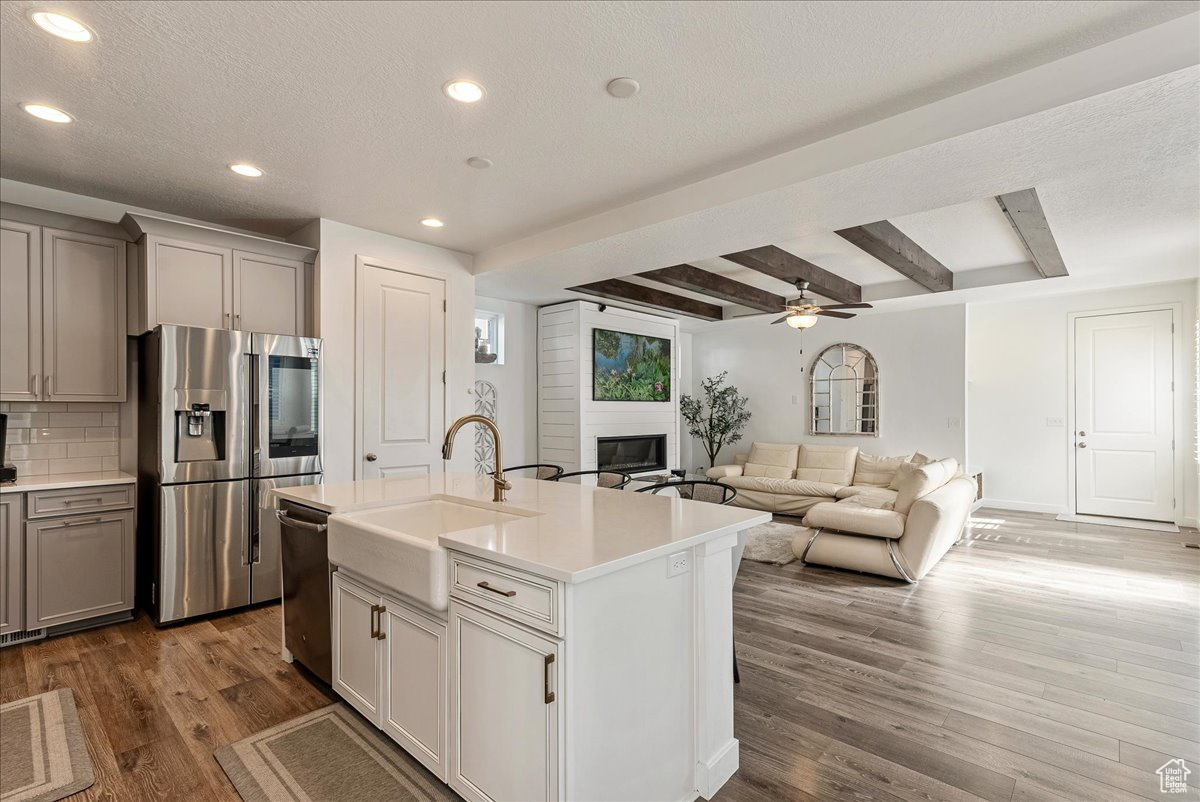 Kitchen featuring beamed ceiling, hardwood / wood-style floors, appliances with stainless steel finishes, a center island with sink, and ceiling fan