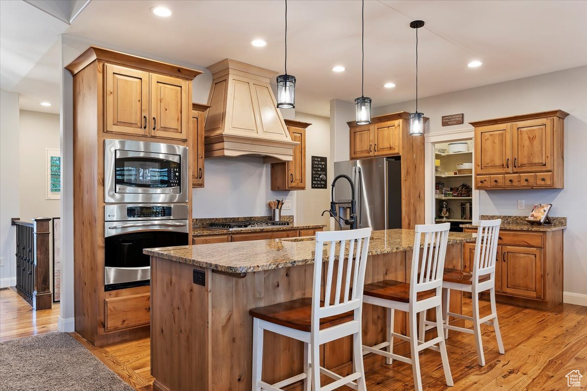 Kitchen with appliances with stainless steel finishes, hardwood flooring, custom range hood, an island with sink, and pendant lighting